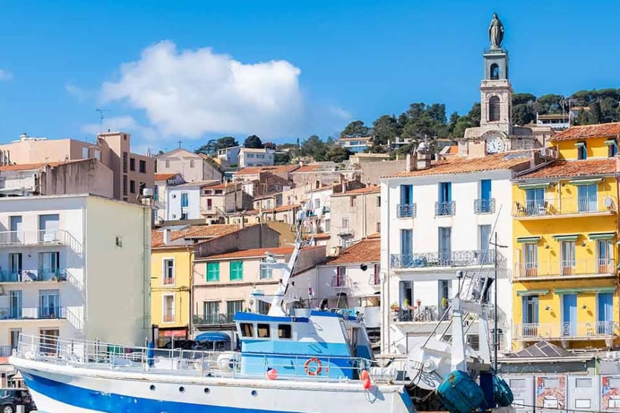 Vue pittoresque d’un port méditerranéen animé, avec un bateau de pêche amarré au premier plan. En arrière-plan, des façades colorées typiques du sud de la France s’élèvent en étages le long de la colline, dominées par un clocher d’église. L’ensemble évoque une atmosphère authentique, conviviale et ensoleillée, idéale pour des projets immobiliers en bord de mer ou dans des quartiers à fort potentiel touristique.