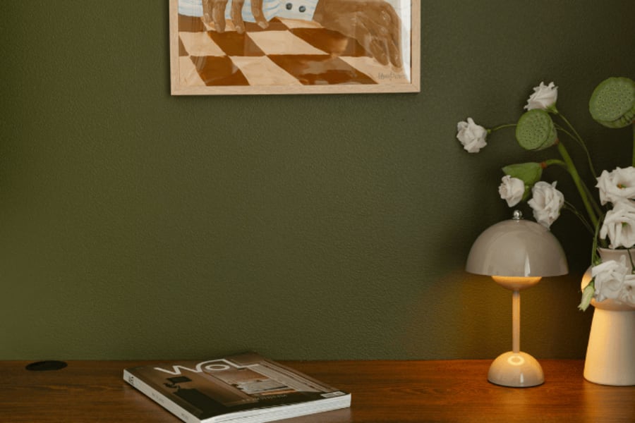 Une table en bois avec un journal et une lampe. Le mur du fond est vert foncé.