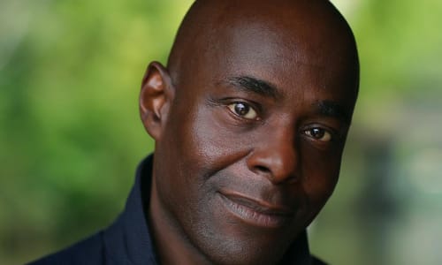 photo of paterson joseph wearing a blue coat with a green leafy background 