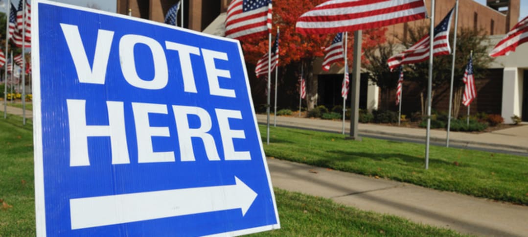 Blue "Vote Here" sign displayed outdoors in front of a sidewalk and multiple American flags staked in the grass.