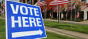 Blue "Vote Here" sign displayed outdoors in front of a sidewalk and multiple American flags staked in the grass.
