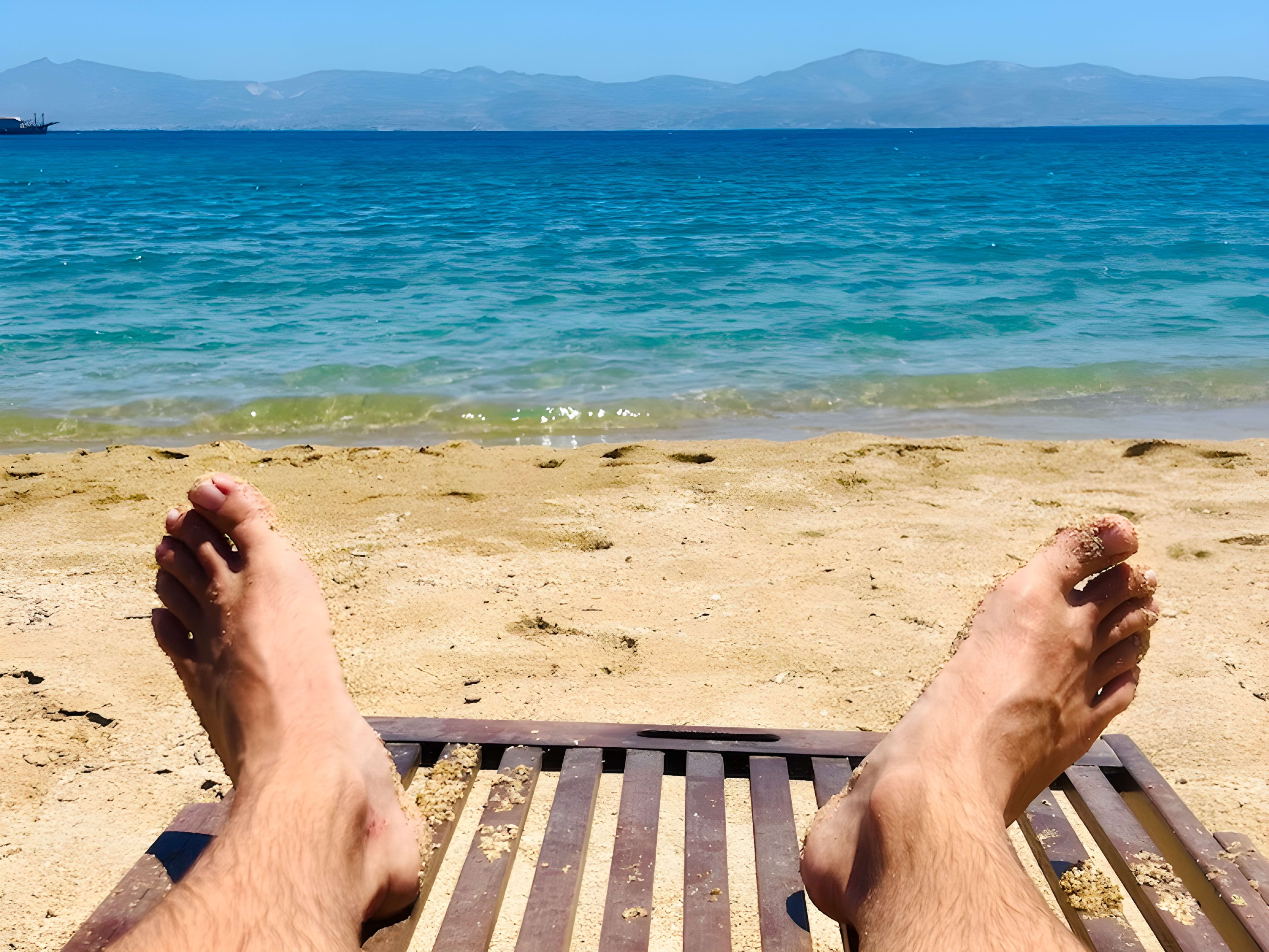 Relaxing on a wooden lounge chair at Logaras Beach in Paros, Greece, with a clear view of the turquoise sea and distant mountains. Beach relaxation is a top activity for those exploring what to do in Paros.