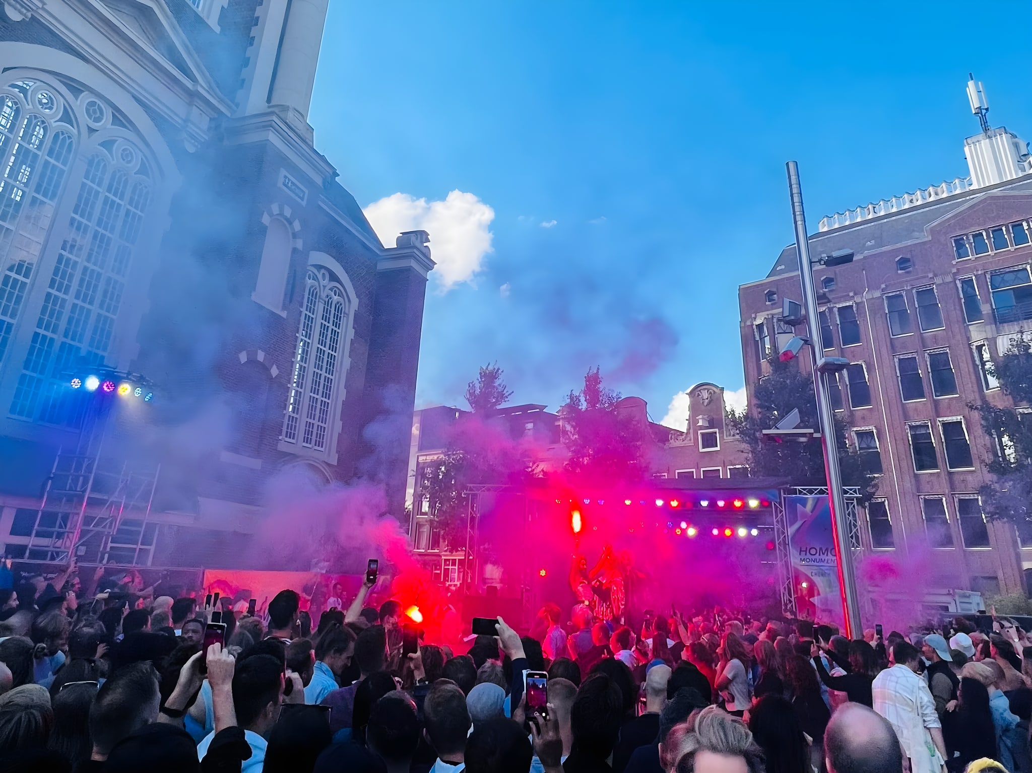 A vibrant crowd at an outdoor concert, illuminated by red stage lights and smoke effects, gathers in front of a historic building under a blue sky—a lively scene reminiscent of Pride festivals. This gay-friendly holiday destination offers an inclusive atmosphere where everyone can celebrate diversity and enjoy the cultural richness and welcoming spirit.