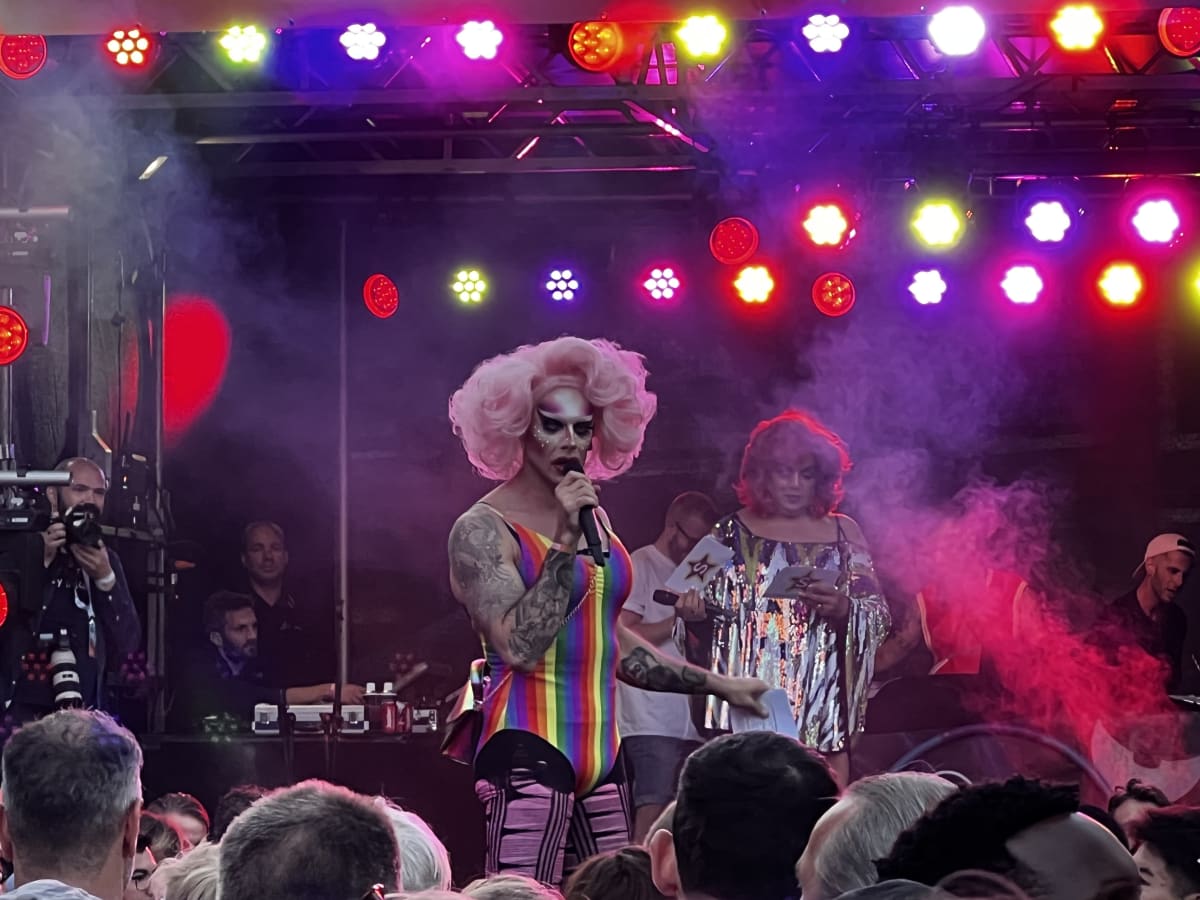 A drag queen with striking pink hair and rainbow attire commands the stage at a gay pride event in the USA, captivating the audience with a powerful performance.