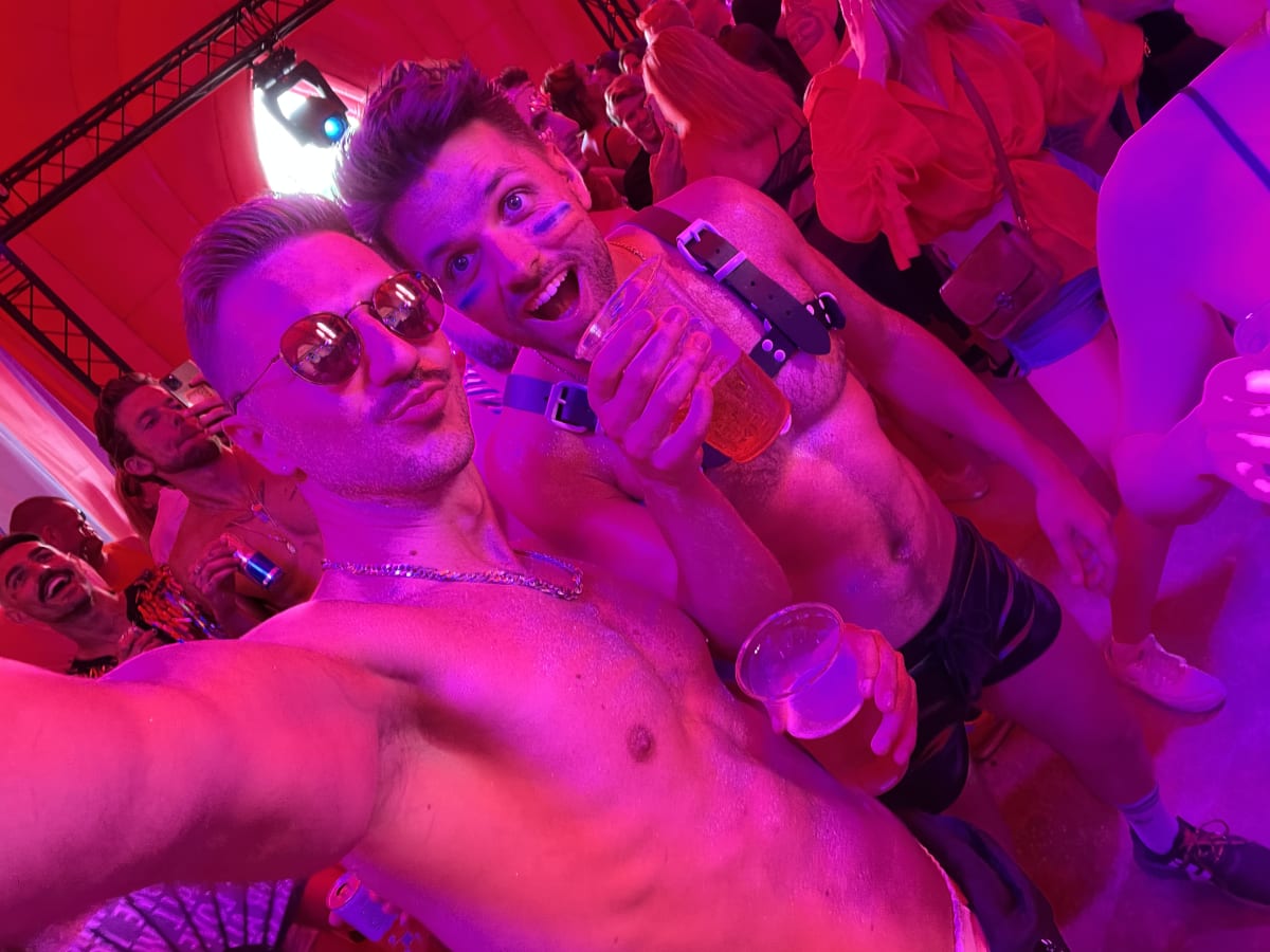 Two shirtless men wearing sunglasses and carrying drinks, exuberantly celebrate gay pride in the USA, surrounded by a crowd in a tent bathed in pink light.