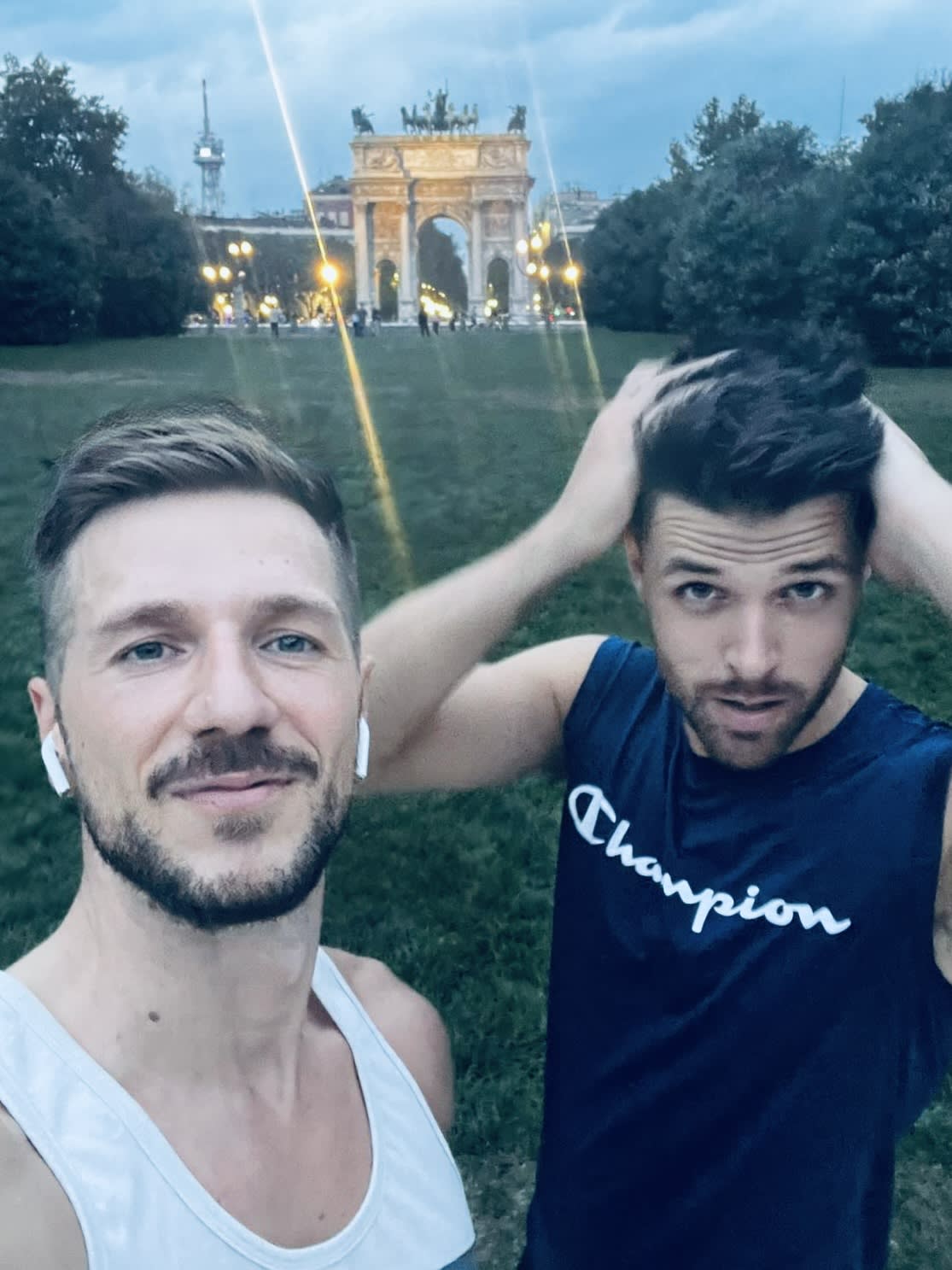 Selfie of two men in casual wear, with the Arco della Pace illuminated in the background during twilight in Milan.