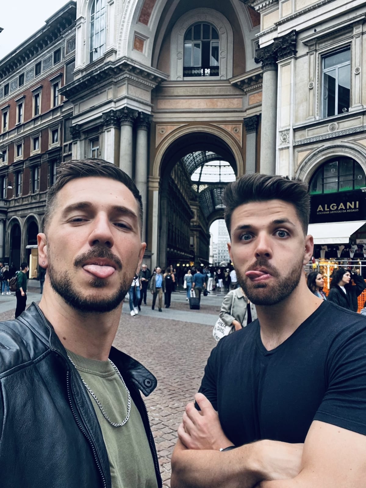 Two men with playful expressions standing in front of the iconic Galleria Vittorio Emanuele II in Milan, surrounded by bustling shoppers.