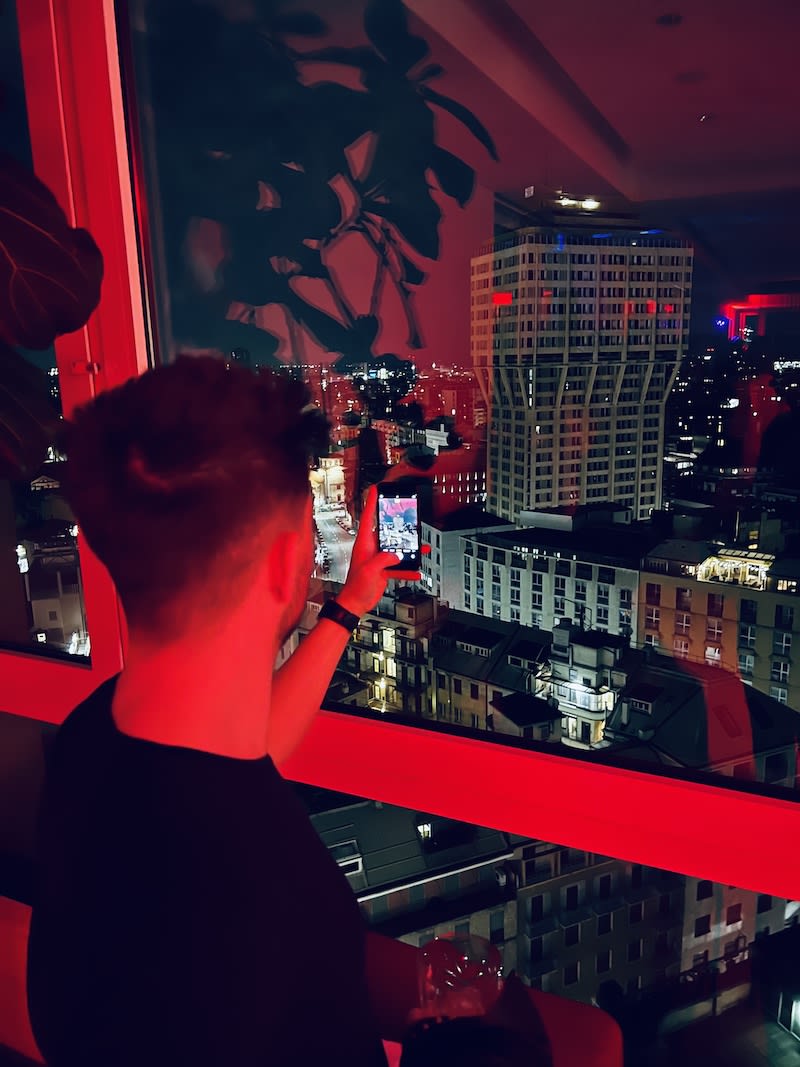 Man capturing the vibrant night cityscape of Milan from a high-rise building with a smartphone.