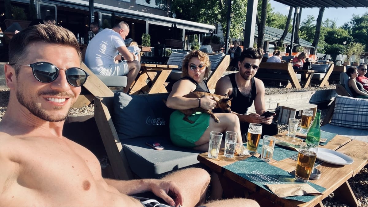 A group of friends relaxing at an outdoor cafe in Belgrade, enjoying drinks and sunny weather. One person is holding a small dog, and the atmosphere is laid-back and cheerful.
