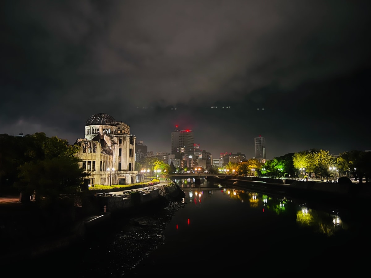 The Hiroshima Peace Memorial, illuminated at night, stands solemnly beside a river with the cityscape of Hiroshima in the background. This poignant site is an essential part of a Honshu Japan itinerary, reminding visitors of the city's history.