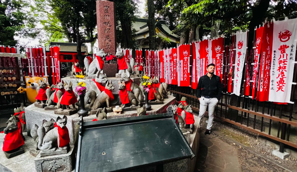 Numerous stone fox statues dressed in red bibs surround a memorial stone at a fox shrine in Tokyo, Japan, with a man standing in the background. Visiting such unique shrines is a memorable part of a Honshu Japan itinerary.