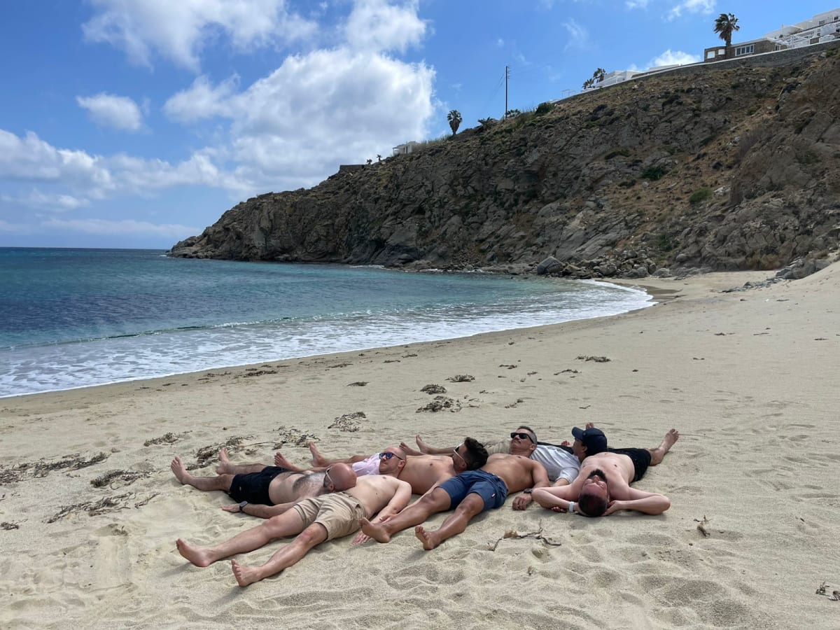 Group of men lying on a sandy Mykonos beach with clear blue water and rocky cliffs in the background.