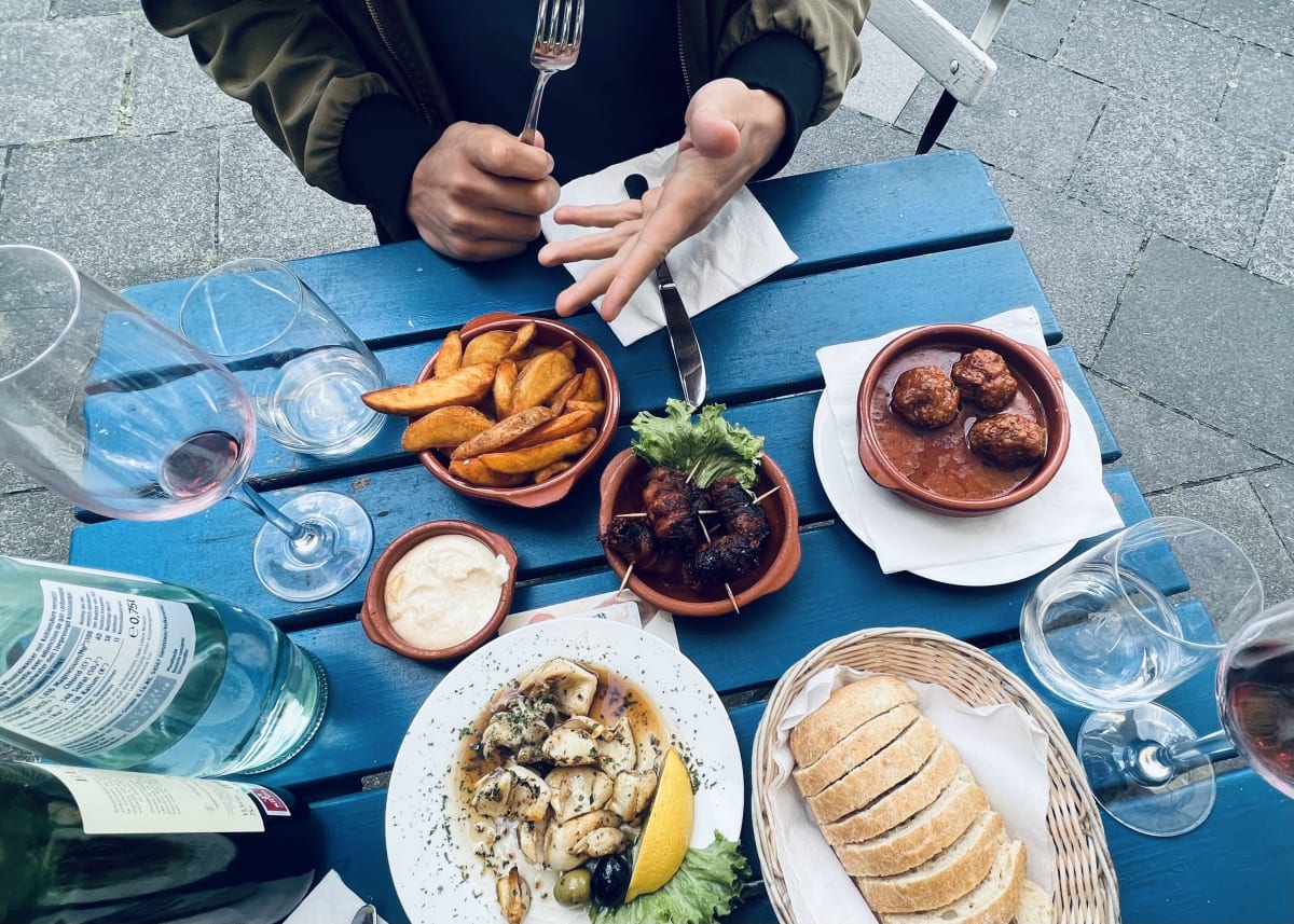 Spanish-style tapas featuring squid, potato wedges, dates wrapped in bacon, meatballs, and bread. Glasses of wine on the side.