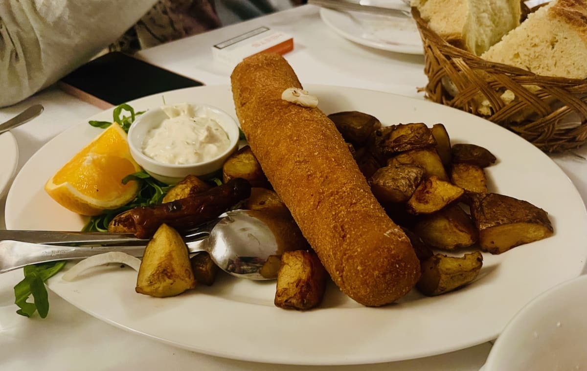 Karađorđeva šnicla, a traditional Serbian food, served on a white plate with roasted potatoes, a slice of orange, and a dipping sauce. The dish consists of a breaded and fried meat roll, garnished with a small dollop of butter.