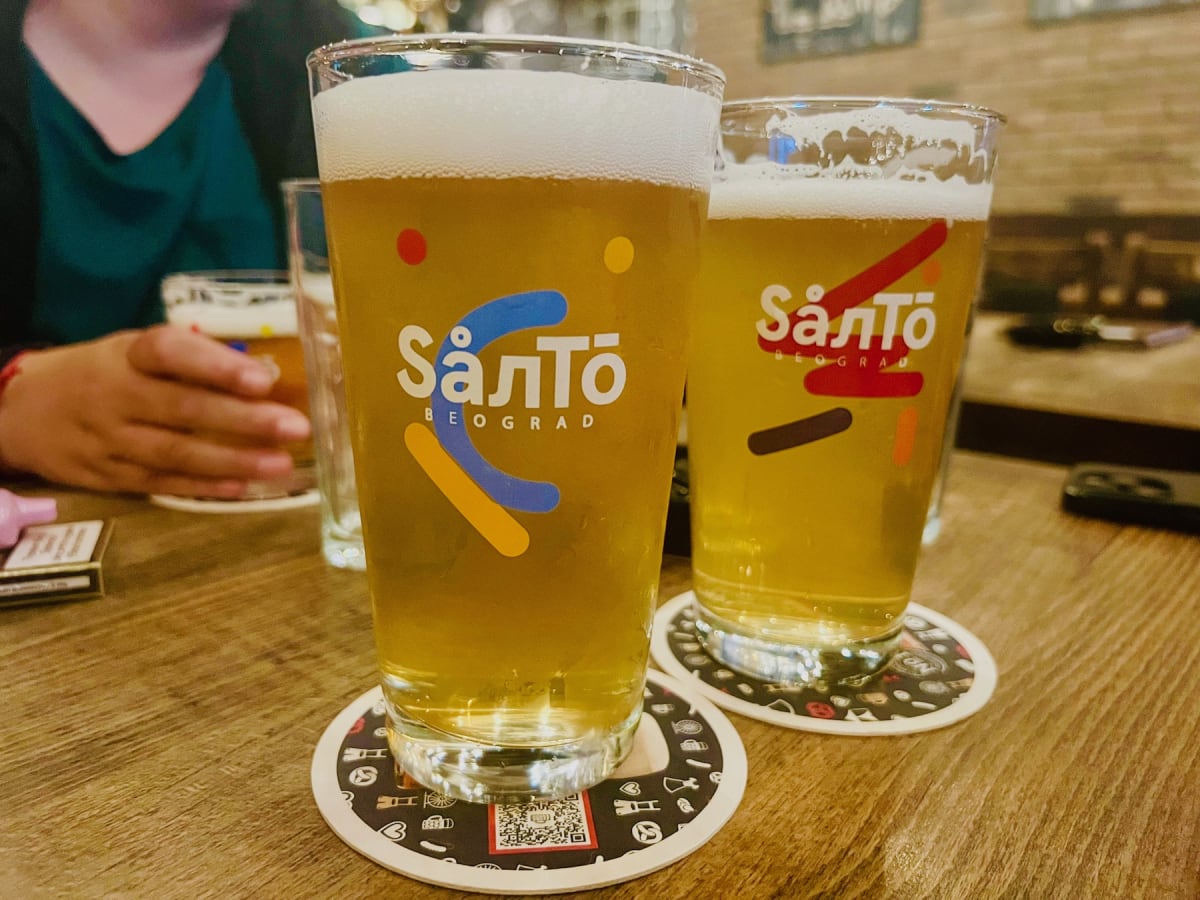 Two glasses of Salto beer from Belgrade, a popular beverage enjoyed with traditional Serbian food, placed on a wooden table with coasters. The beer glasses are half-full, with foam at the top, and people in the background enjoying their drinks.