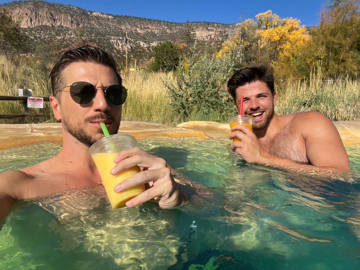 Two men enjoy drinks while relaxing in a hot spring with mountains in the background. Their smiles and the beautiful setting are perfect to brighten your day.