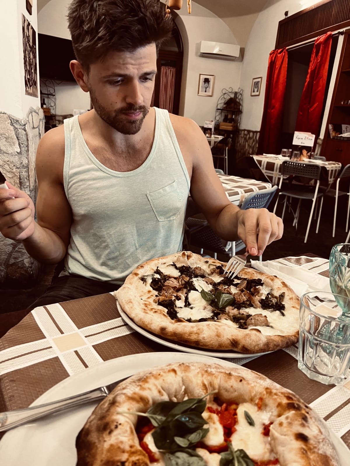 A man in a tank top enjoys a Neapolitan pizza at a cozy restaurant in Naples, Italy, highlighting the city’s culinary delights. This moment captures the casual dining experience in gay Naples, Italy.