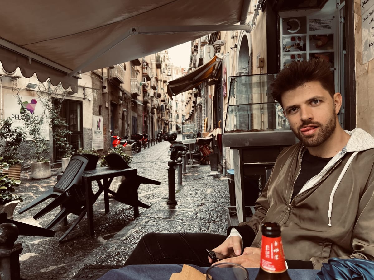 A man relaxes at an outdoor cafe in a narrow, cobblestone street in Naples, Italy, enjoying the local ambiance. This scene captures the laid-back atmosphere of gay Naples, Italy.