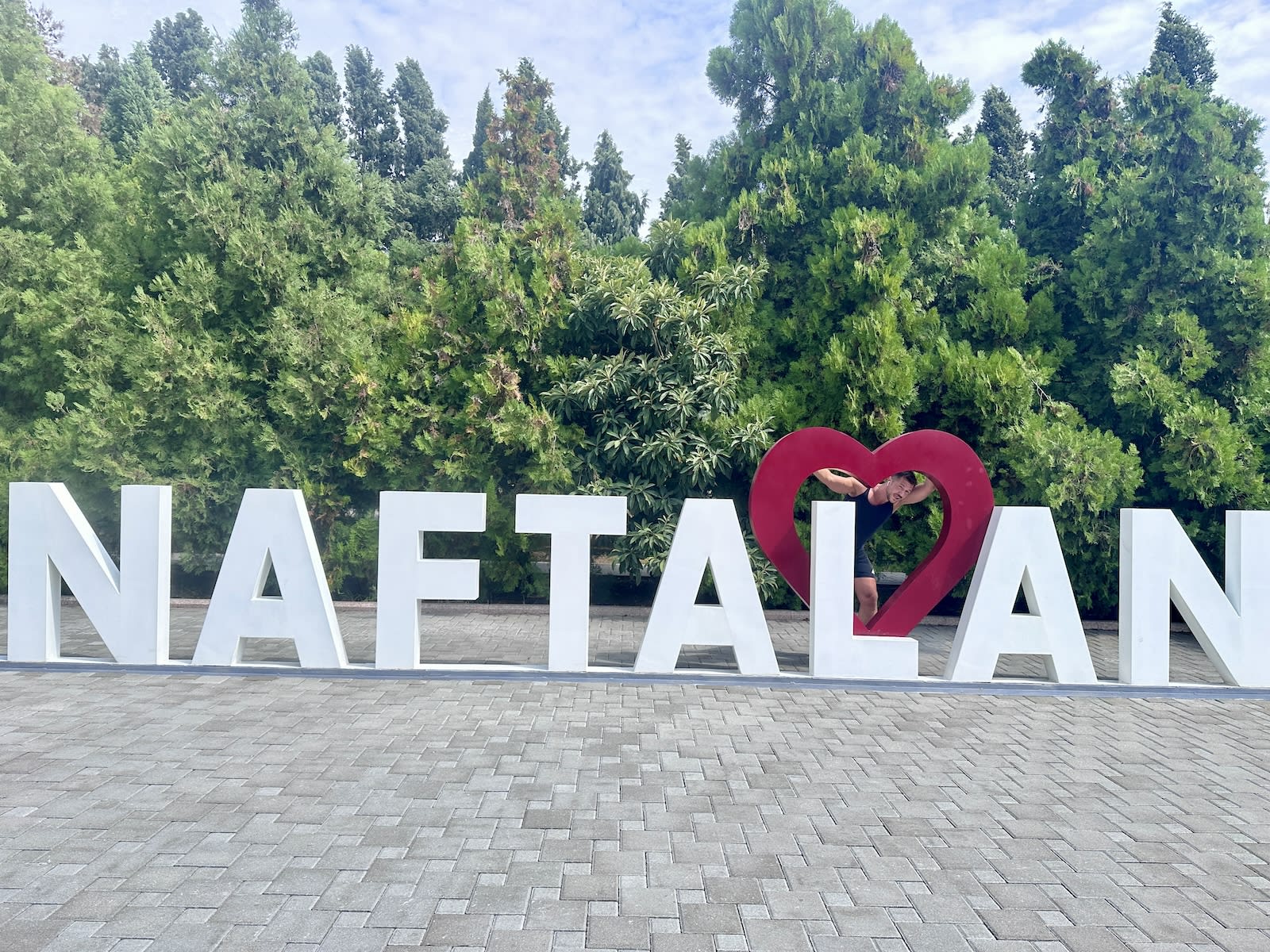 Large 'NAFTALAN' sign with a heart-shaped hole and a person peeking through it, against greenery.