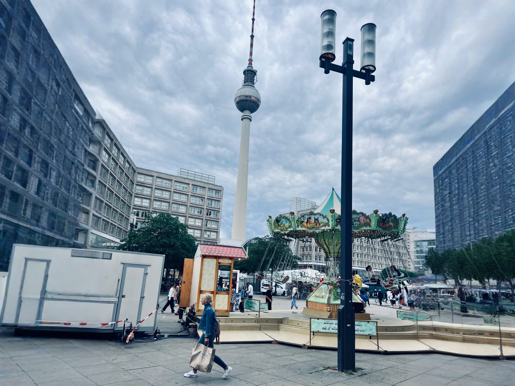 Daily life in Alexanderplatz with the Berlin TV Tower, or Fernsehturm, towering in the background. Foreground activity includes pedestrians, a carousel, and street lamps, capturing the blend of urban life and tourist attractions in central Berlin.