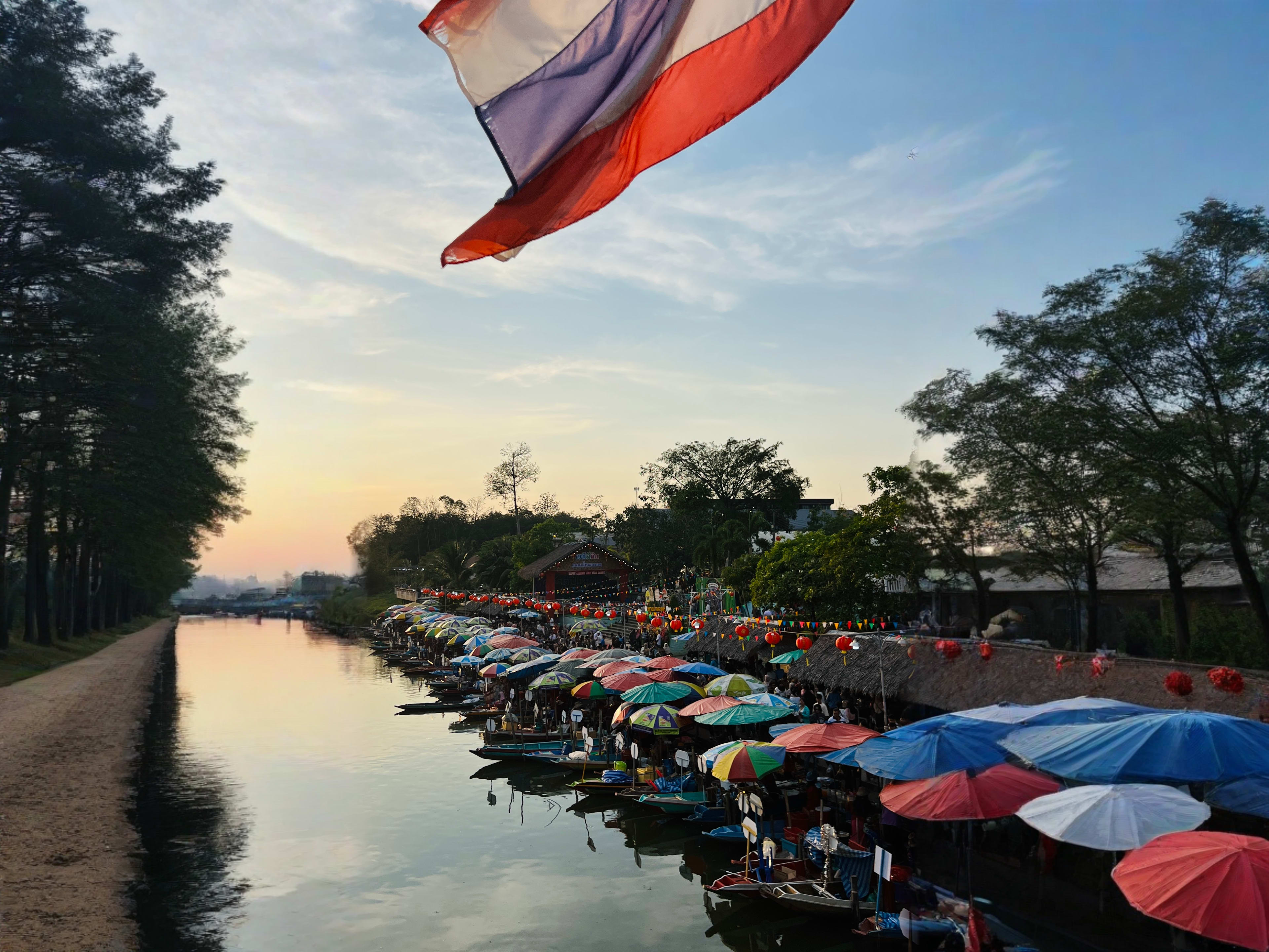 Colorful umbrellas cover boats at a lively riverside market at sunset in Hat Yai, Thailand—a hidden gem with vibrant stalls, fluttering flags, and lush trees along the riverbank; this iconic scene is a top highlight among things to do Hat Yai for visitors seeking authentic local experiences.
