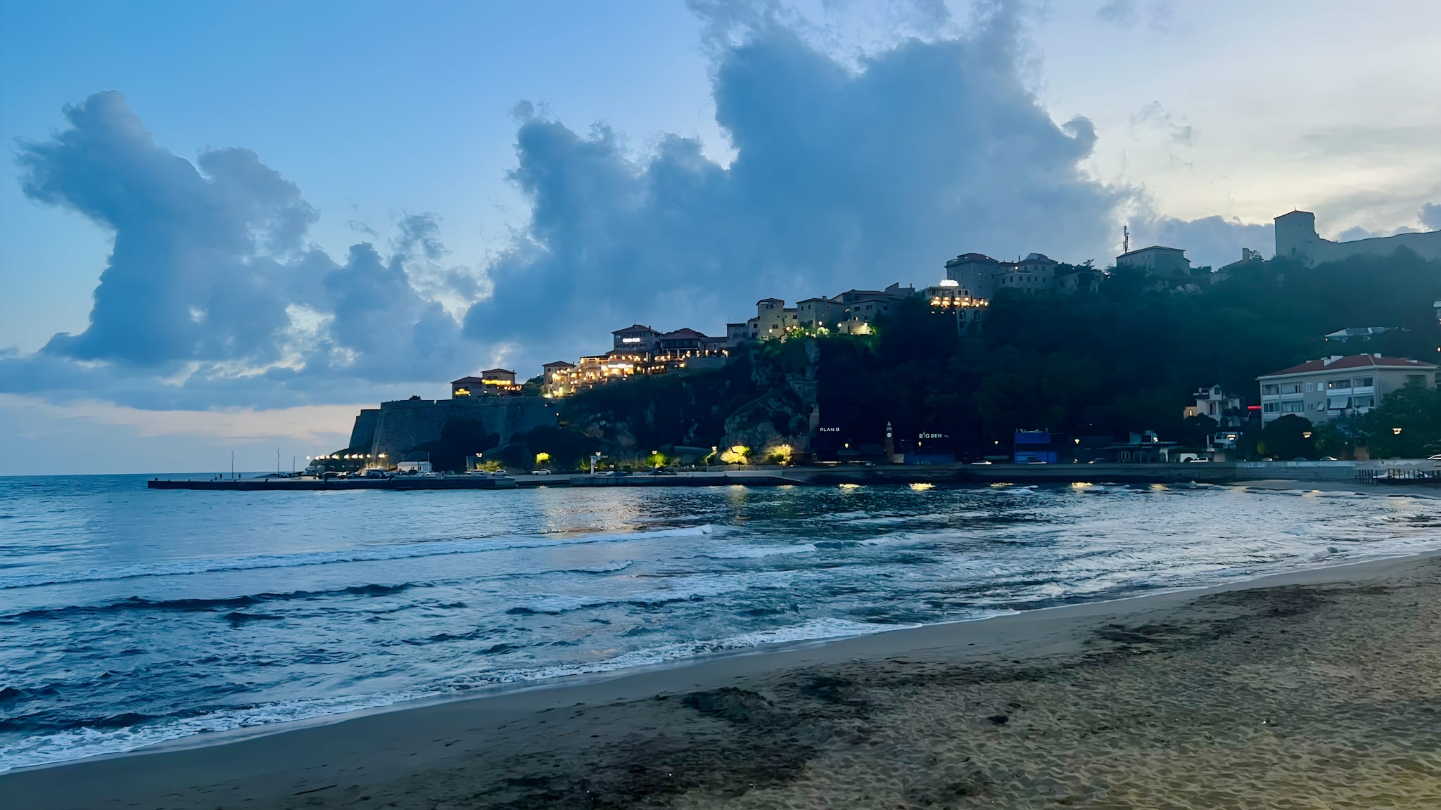 Beach at dusk with gentle waves lapping the sandy shore in the foreground. Illuminated buildings dot a distant hill beneath a cloudy sky, creating an enchanting scene perfect for a coastal town stop on your Montenegro itinerary.