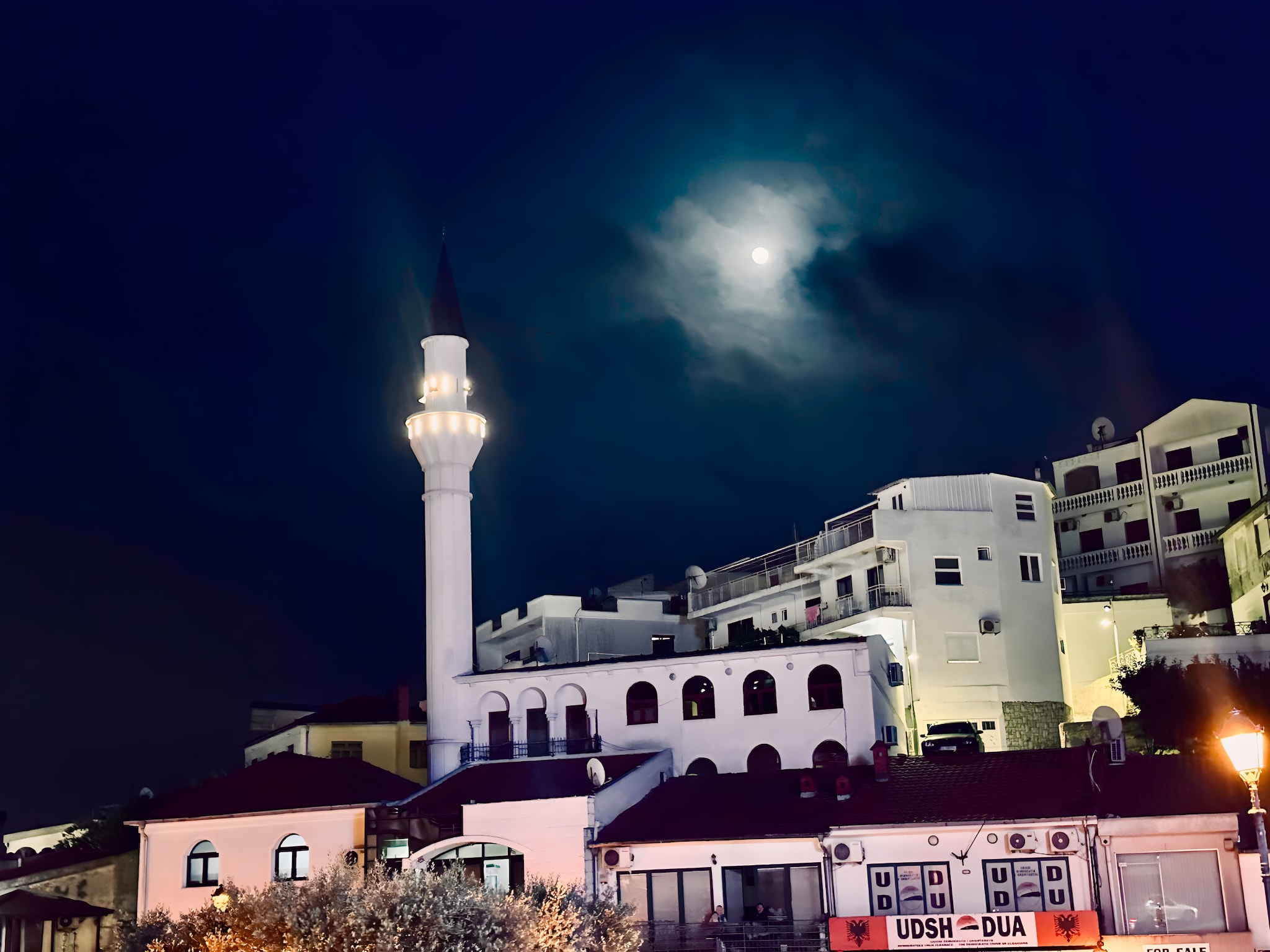 A lit mosque minaret rises gracefully amid a cluster of buildings under a cloudy night sky, where the moon peeks through. It's an enchanting sight to include in your Montenegro itinerary, bringing the region's rich cultural tapestry to life.