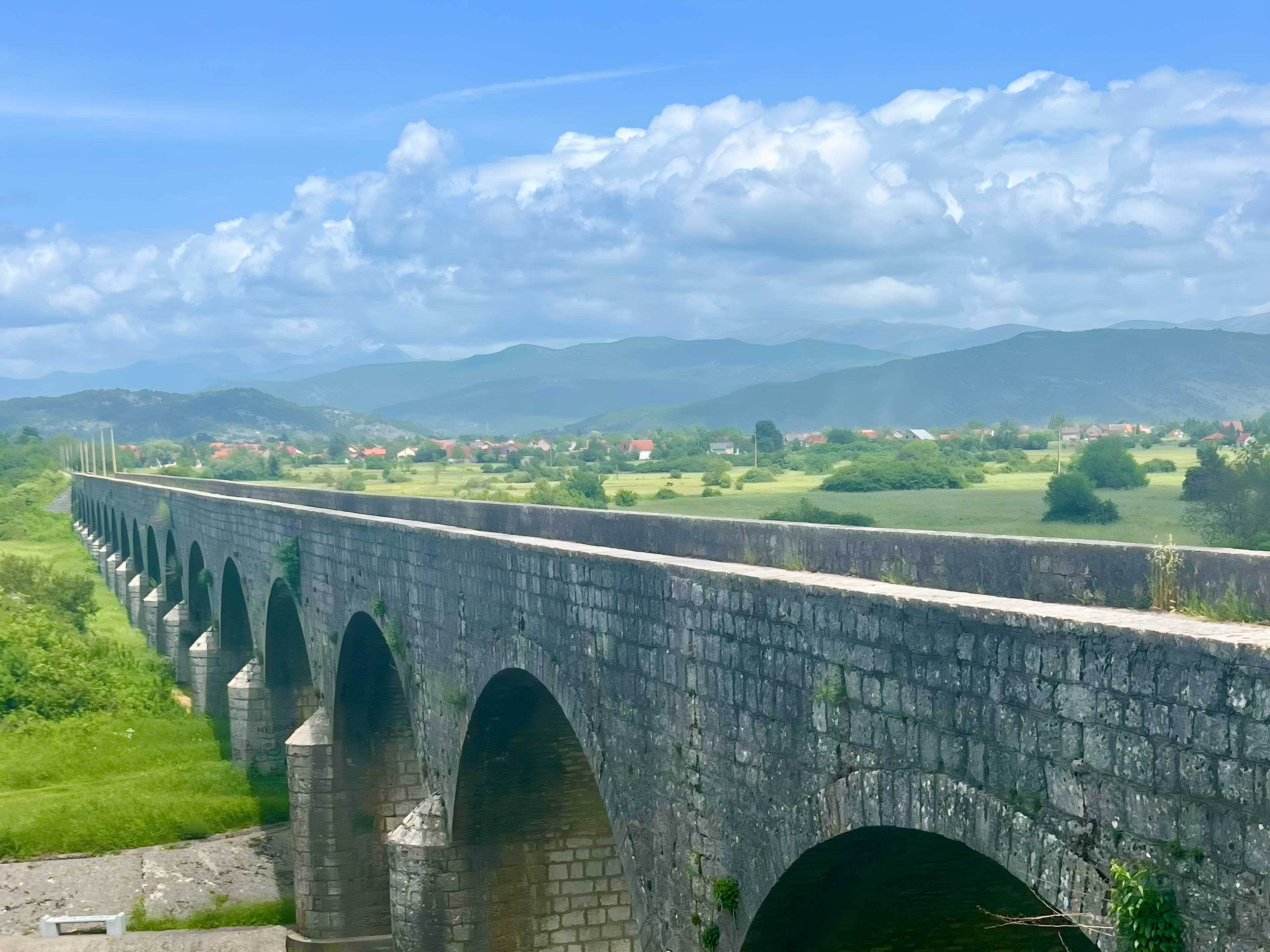 A long stone bridge with multiple arches gracefully spans a grassy landscape, providing a picturesque start to any Montenegro itinerary. Distant mountains and scattered houses adorn the horizon under a partly cloudy sky, setting the stage for an authentic Balkan adventure.