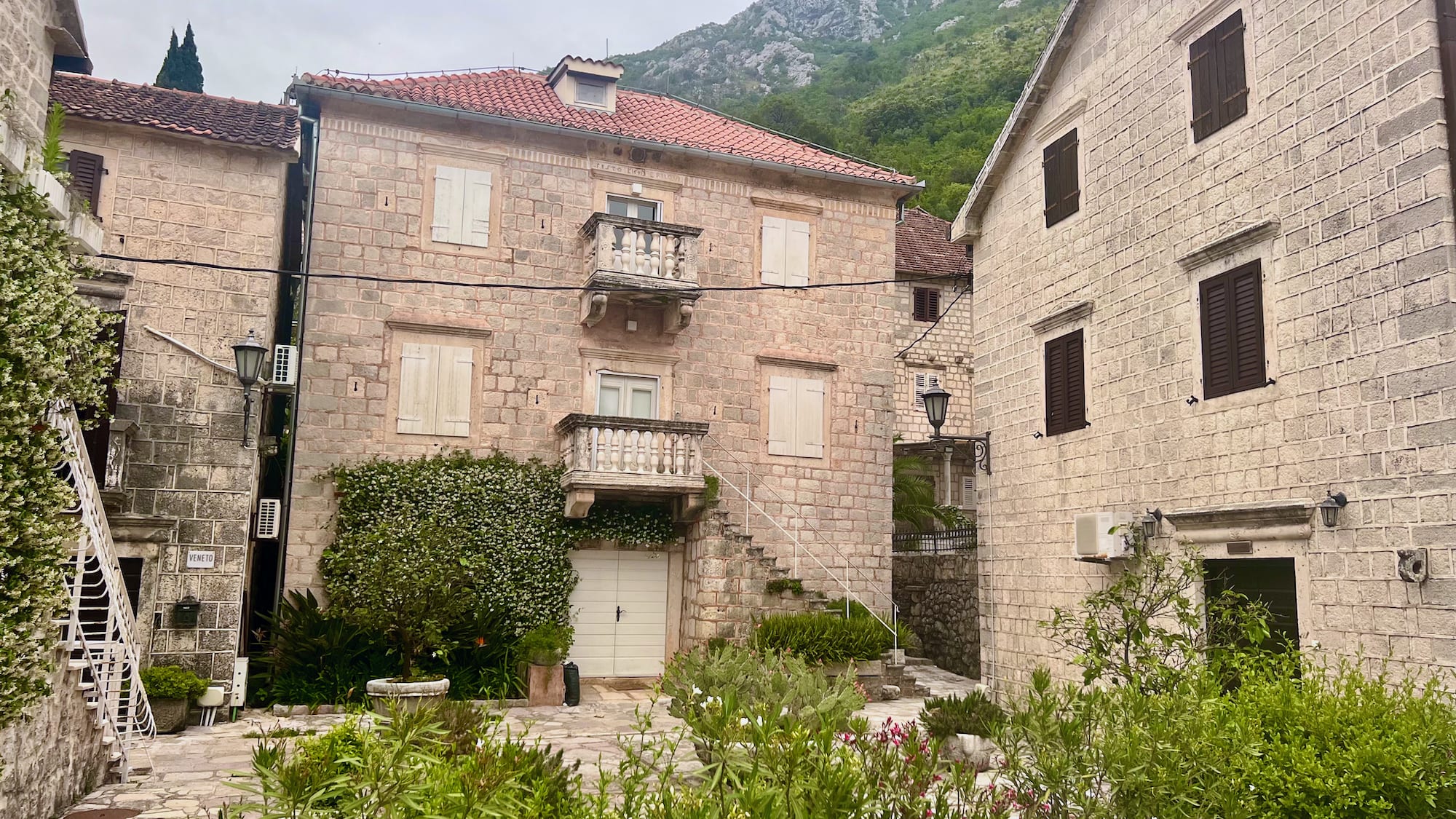 A stone courtyard surrounded by rustic buildings adorned with balconies and lush greenery creates a charming stop on your Montenegro itinerary, adding character to your journey. Nestled among scenic mountains, it captures the essence of Montenegro's coastal towns, offering a glimpse into the enchanting ambiance of this unforgettable Balkan adventure.