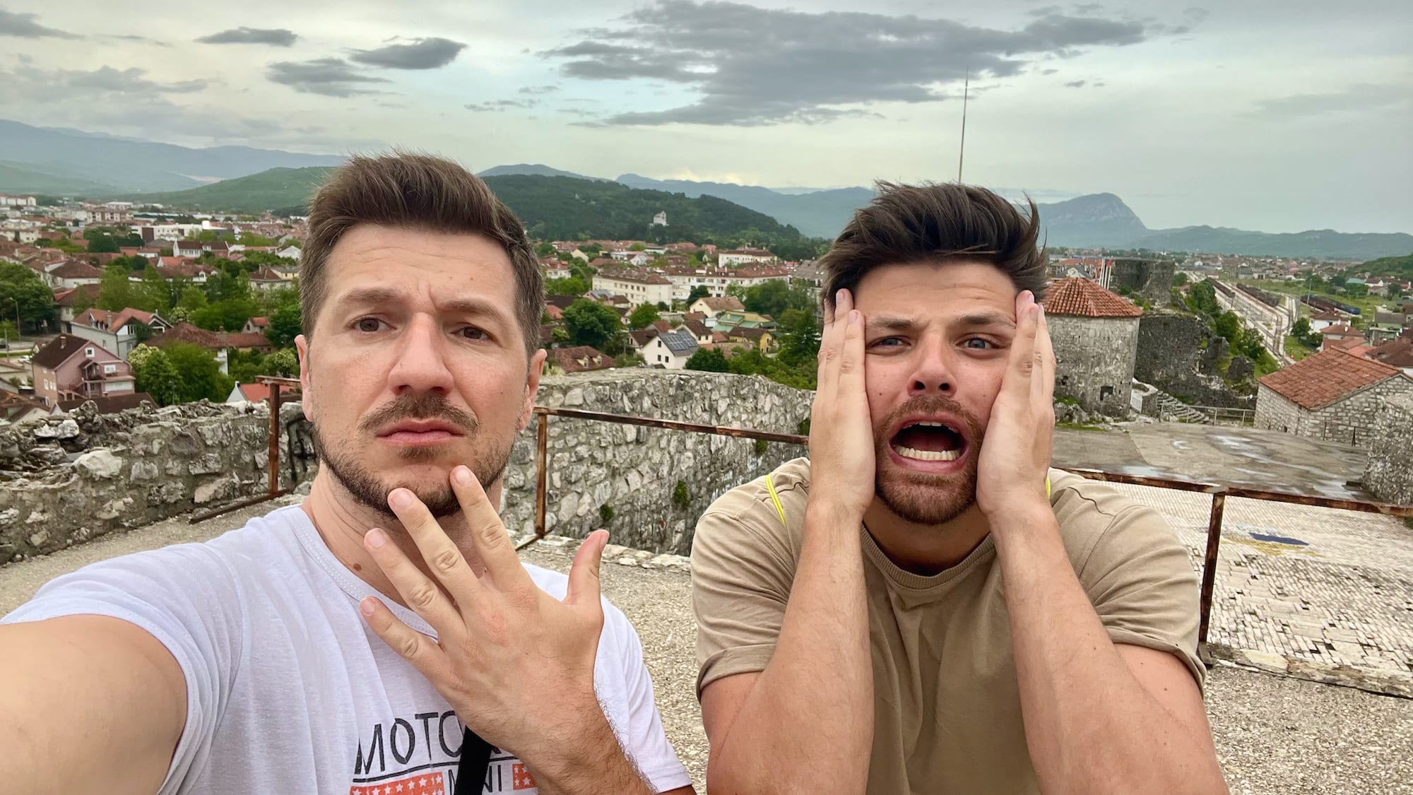 Two men outdoors, one with hands on face and the other touching his chin, showing surprise amidst their Balkan adventure. A panoramic view of a coastal town backed by rolling hills unfolds behind them, capturing the essence of their Montenegro itinerary as they explore this scenic destination.