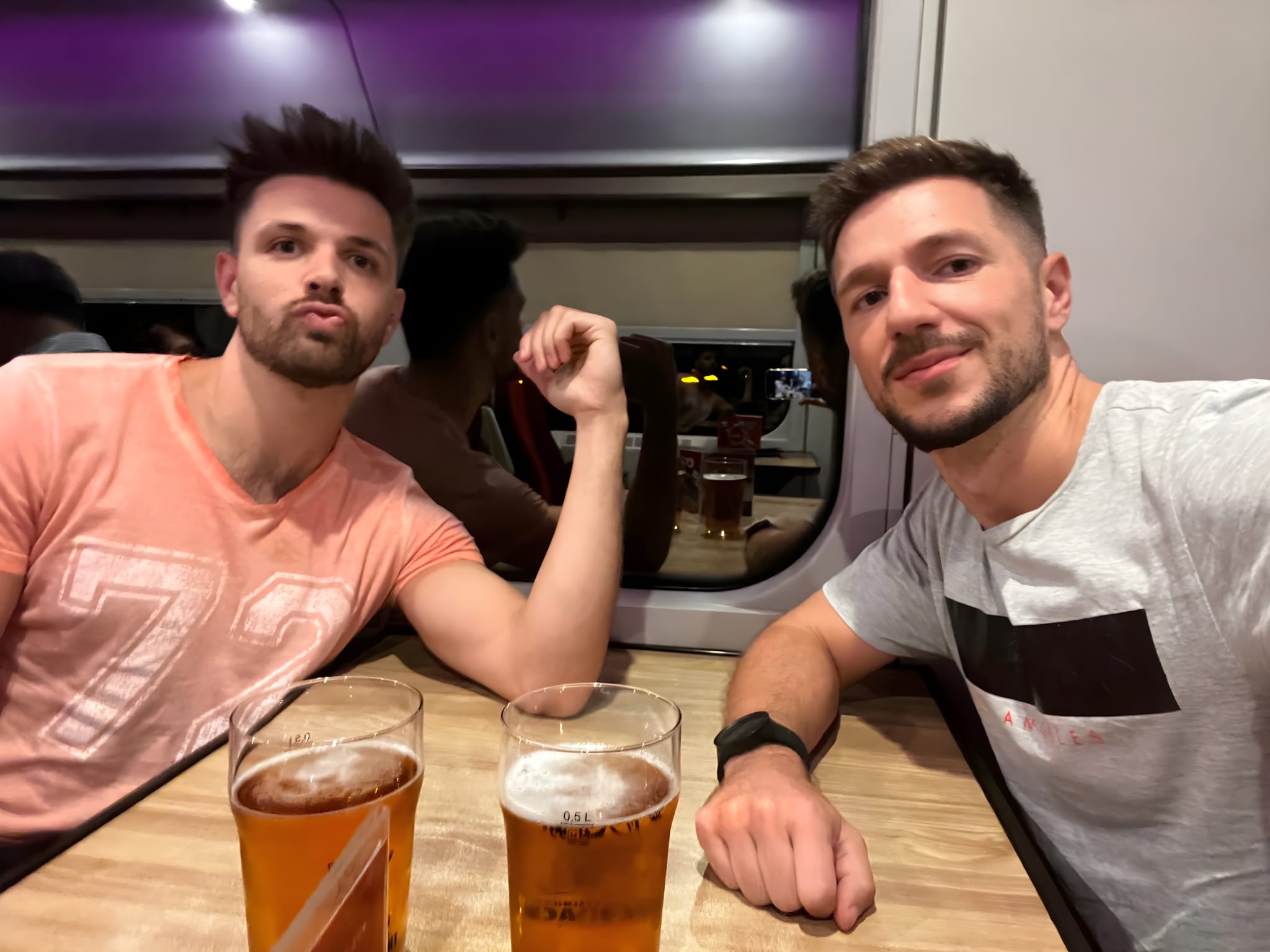 Two people sit at a table on a train, pint glasses of beer before them. One makes a playful face while the other smiles, enjoying this epic way to travel.
