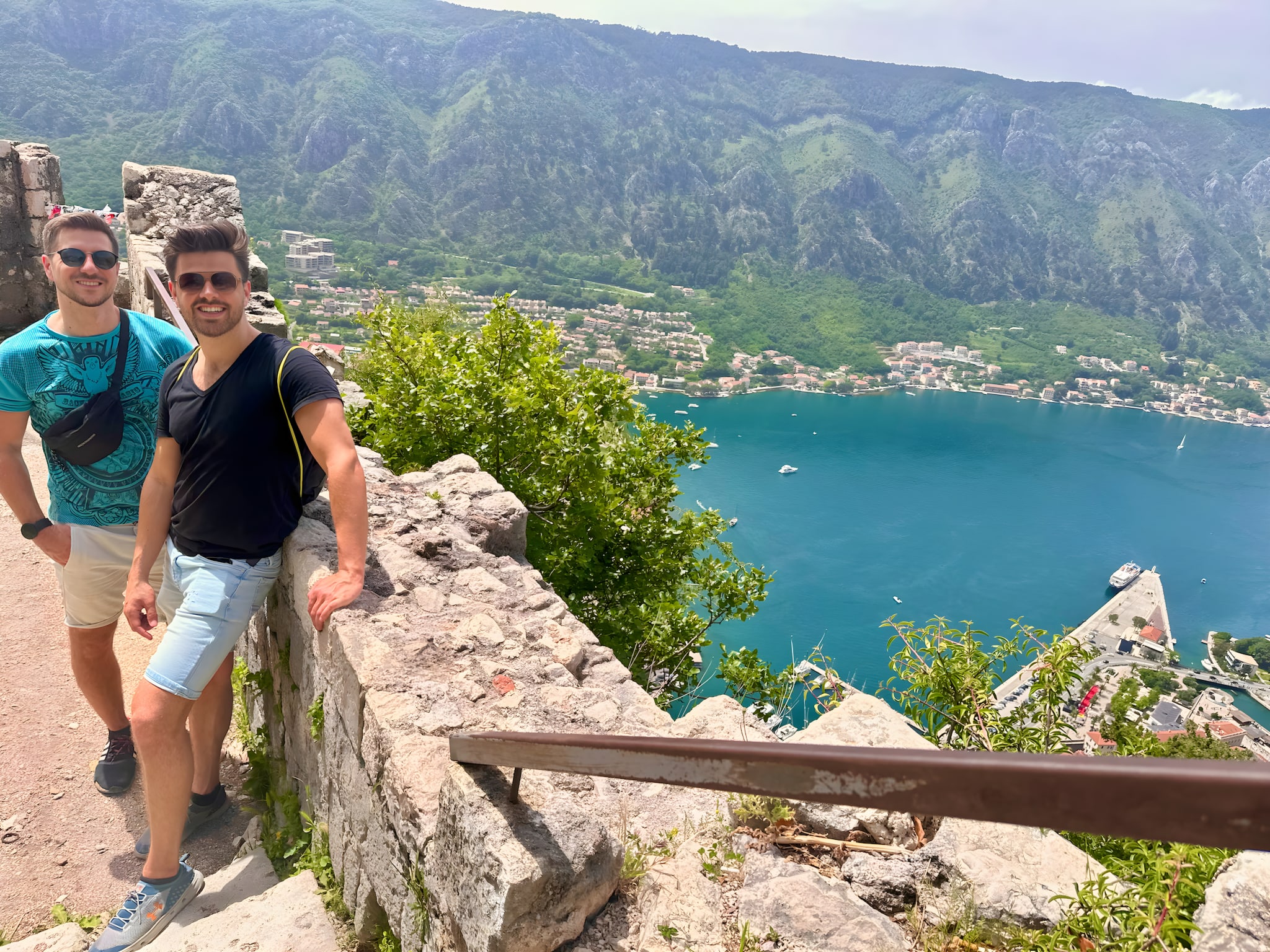 Two people stand by a stone wall overlooking a stunning mountainous landscape with a picturesque bay. As part of their Montenegro itinerary, the view includes lush greenery, a charming village, and the shimmering water below—a perfect stop on any Balkan adventure.
