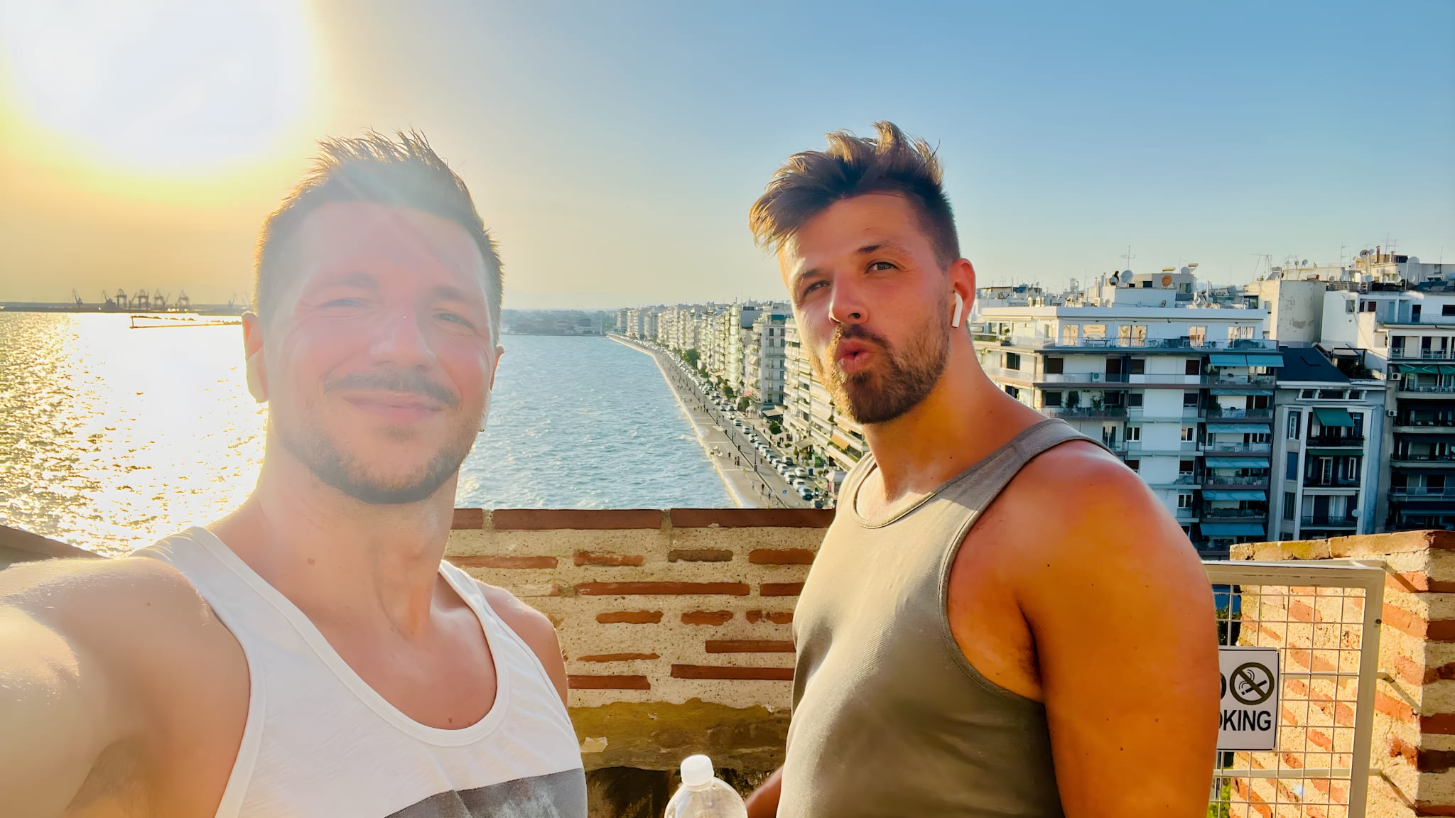 Two men take a selfie on a sunny day, celebrating in a vibrant waterfront cityscape renowned as a gay-friendly holiday destination. With a distant port in the background and one holding a water bottle, their joy captures the essence of pride festivals found here, showcasing why this lively locale is favored by LGBTQ+ travelers seeking inclusive and welcoming experiences.