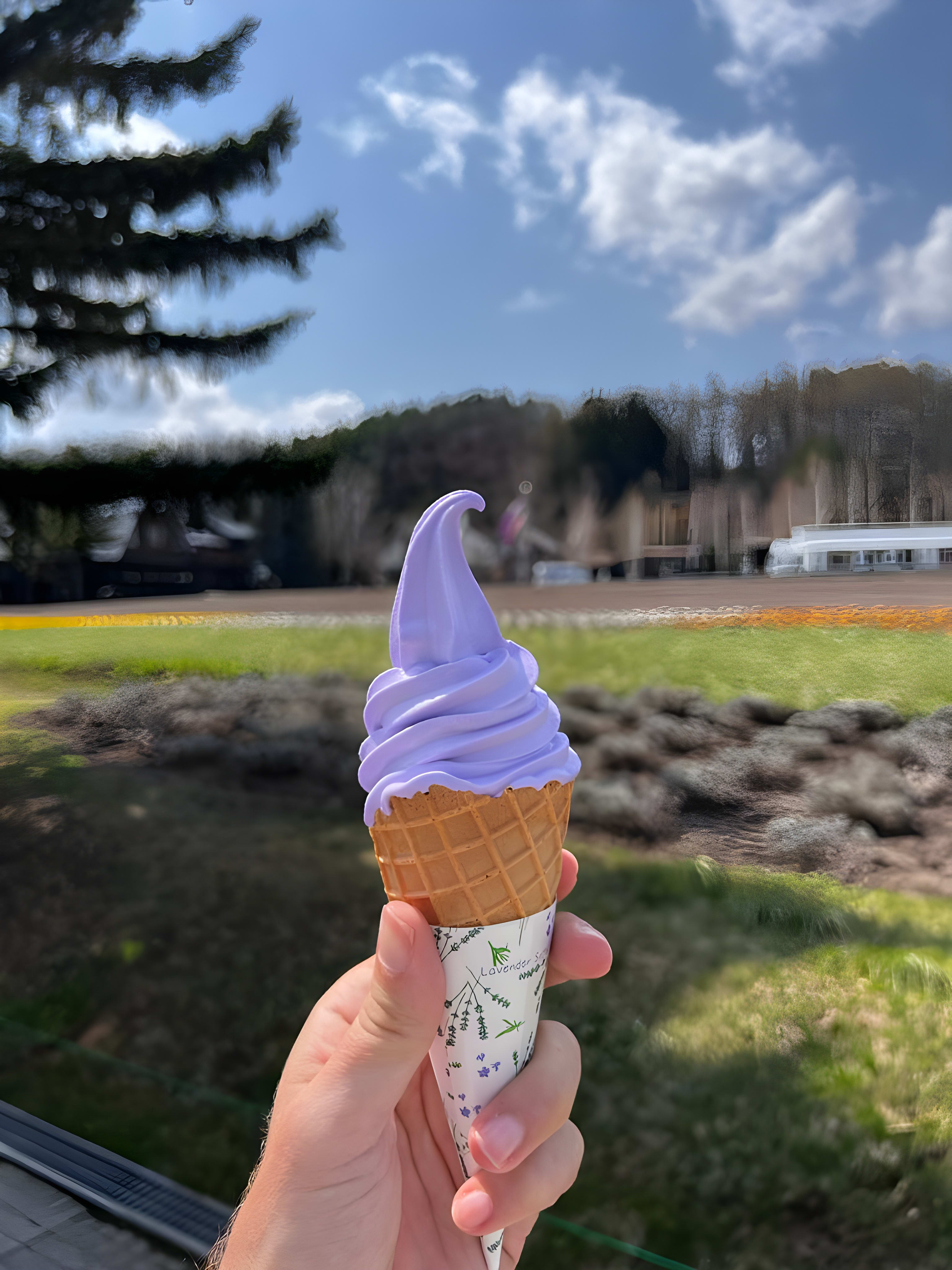 A hand holds a creamy lavender soft serve ice cream cone outdoors against a backdrop of lush grass, leafy trees, and a bright blue sky with fluffy clouds. This delicious treat is perfect for cooling off and adds a sweet moment to any japan itinerary or travel adventure in Japan’s scenic countryside.