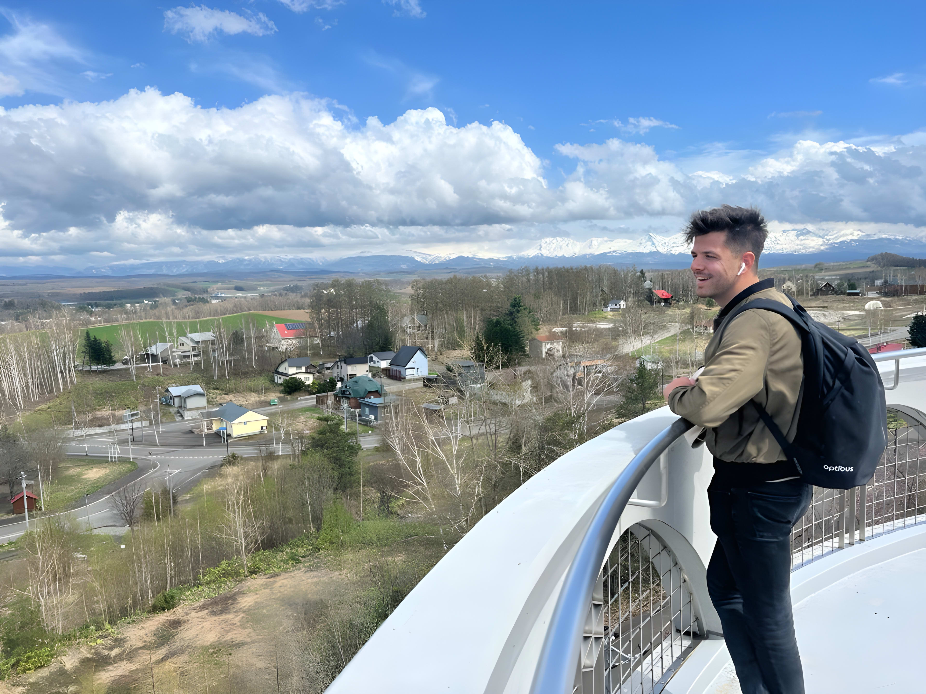 A man with a backpack stands on a curved observation deck in Hokkaido, an ideal spot for any japan itinerary, gazing over a rural landscape dotted with houses and trees. Snow-capped mountains rise in the distance beneath a partly cloudy sky, creating a picturesque travel moment.