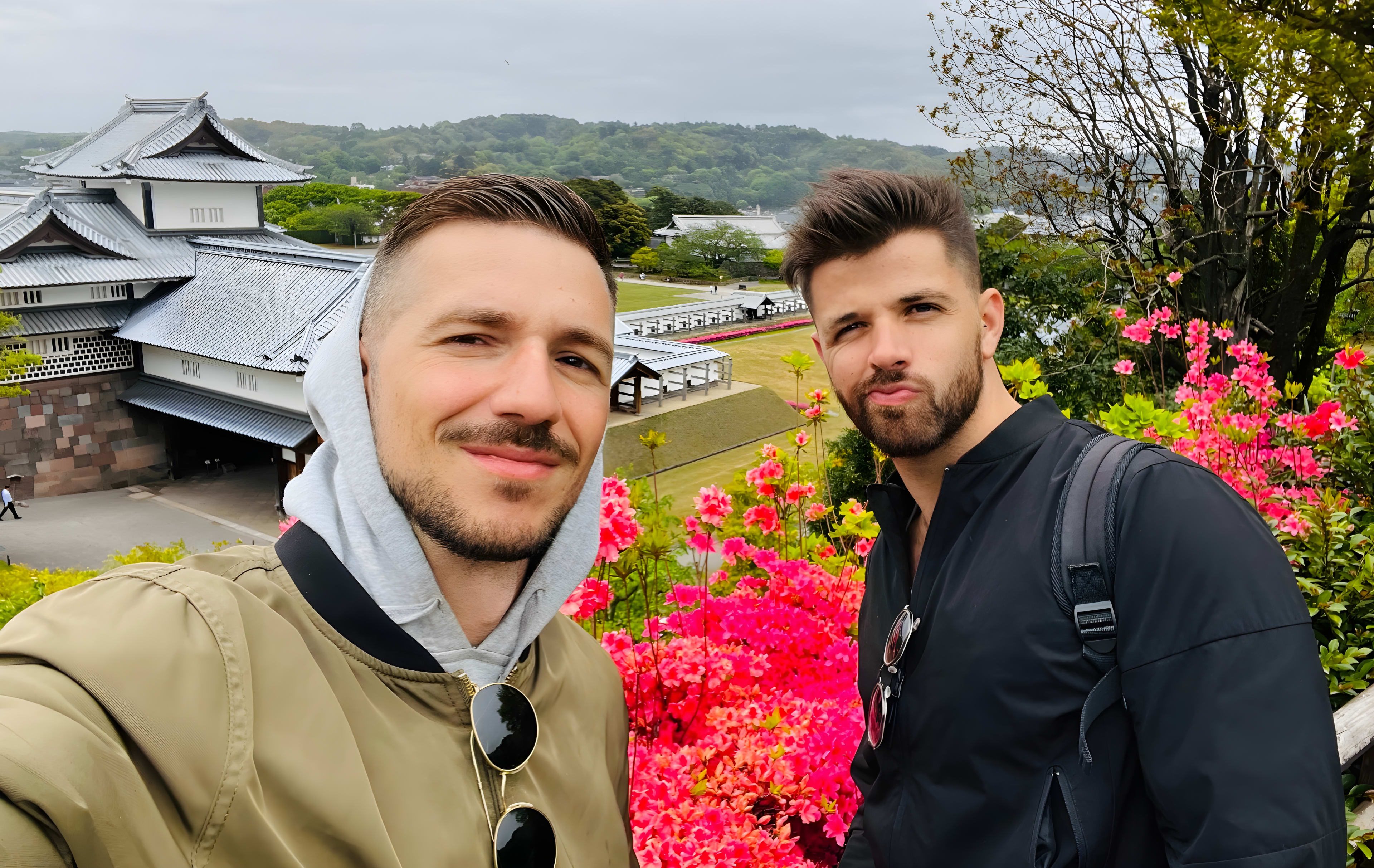 Two men smiling for a selfie in front of a traditional Japanese building, surrounded by vibrant pink flowers and lush greenery on a cloudy day—an unforgettable memory from their Japan itinerary and cultural travel adventure.