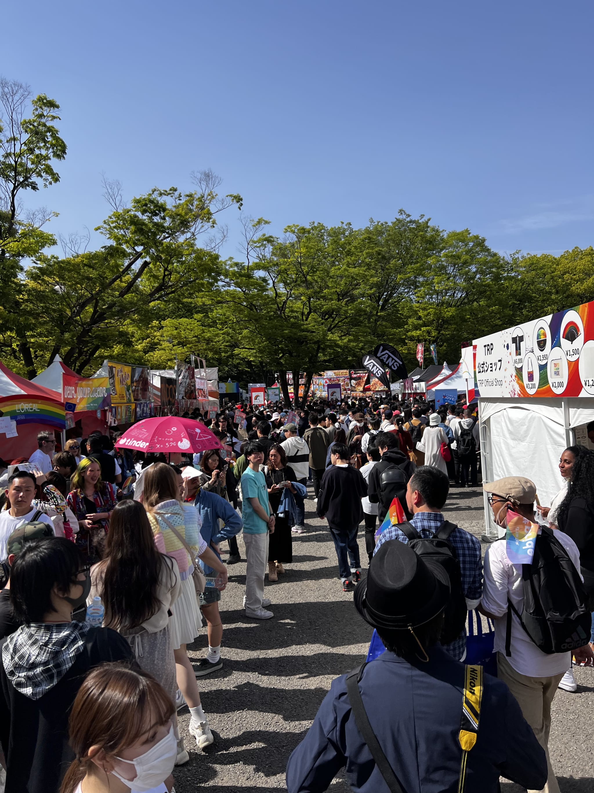 A bustling outdoor festival unfolds under clear skies, where diverse attendees meander among colorful tents and stalls. People proudly hold rainbow flags and umbrellas, joyfully celebrating Asian Gay Pride at the Gay Pride Asia 2025 event. The pathways, lushly lined with trees, resonate with a vibrant atmosphere of diversity and joy, epitomizing the spirit of unity and acceptance.