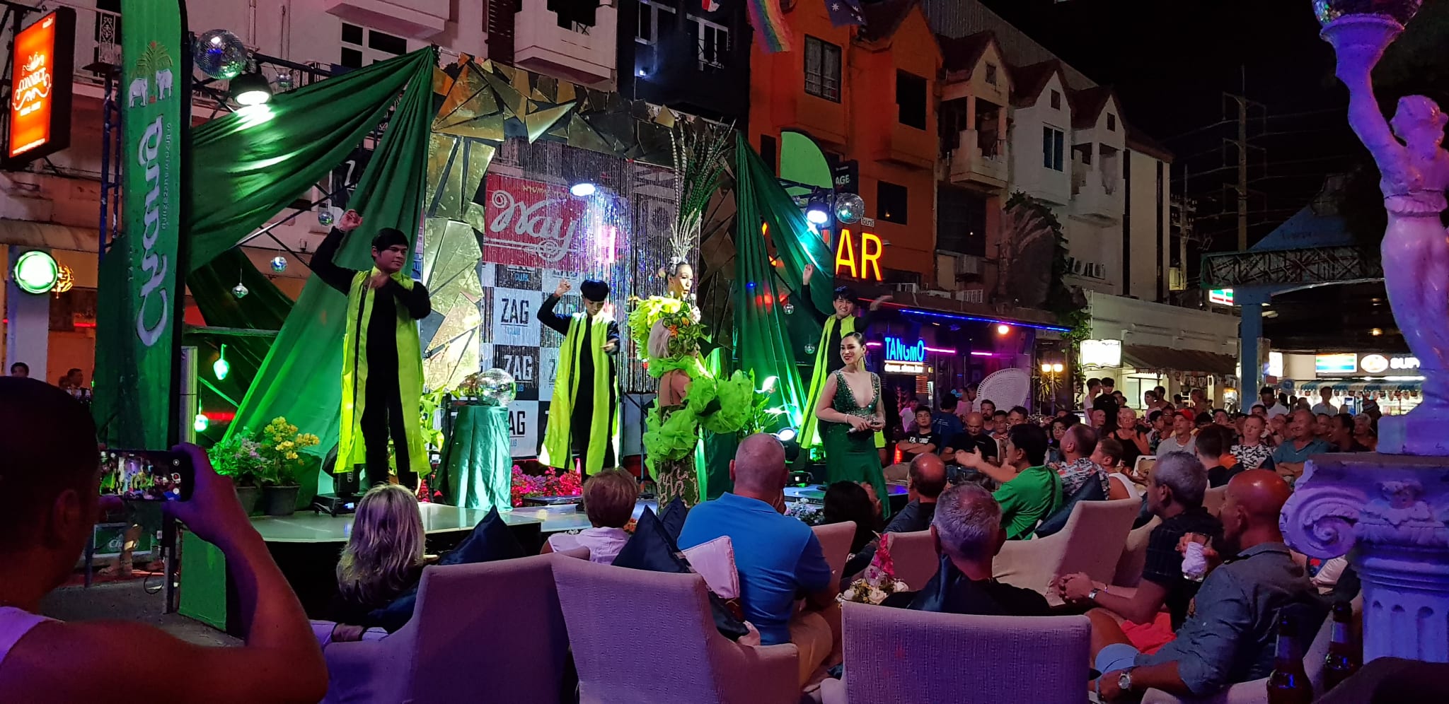 A lively street performance features three people on stage dressed in black and green robes. The audience, celebrating Asian Pride Month and Asia events, sits in chairs, watching intently. Bright green and colorful decorations surround the stage. A mix of people capture the moment with their phones amid vibrant city nightlife, adding to the festive atmosphere.