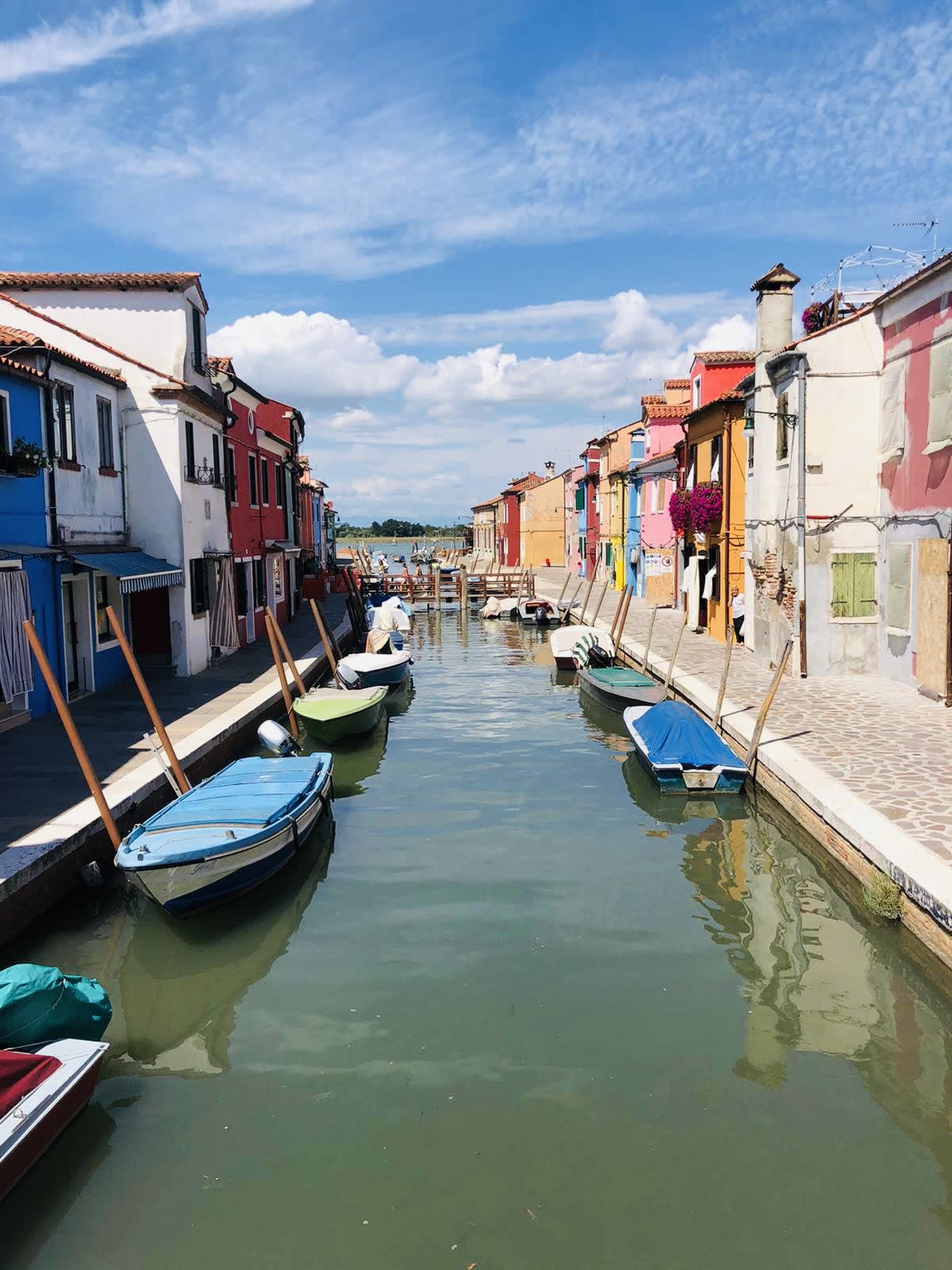 A picturesque view of colorful buildings along a canal with boats moored at the edges under a partly cloudy sky captures the vibrant spirit of Venice.