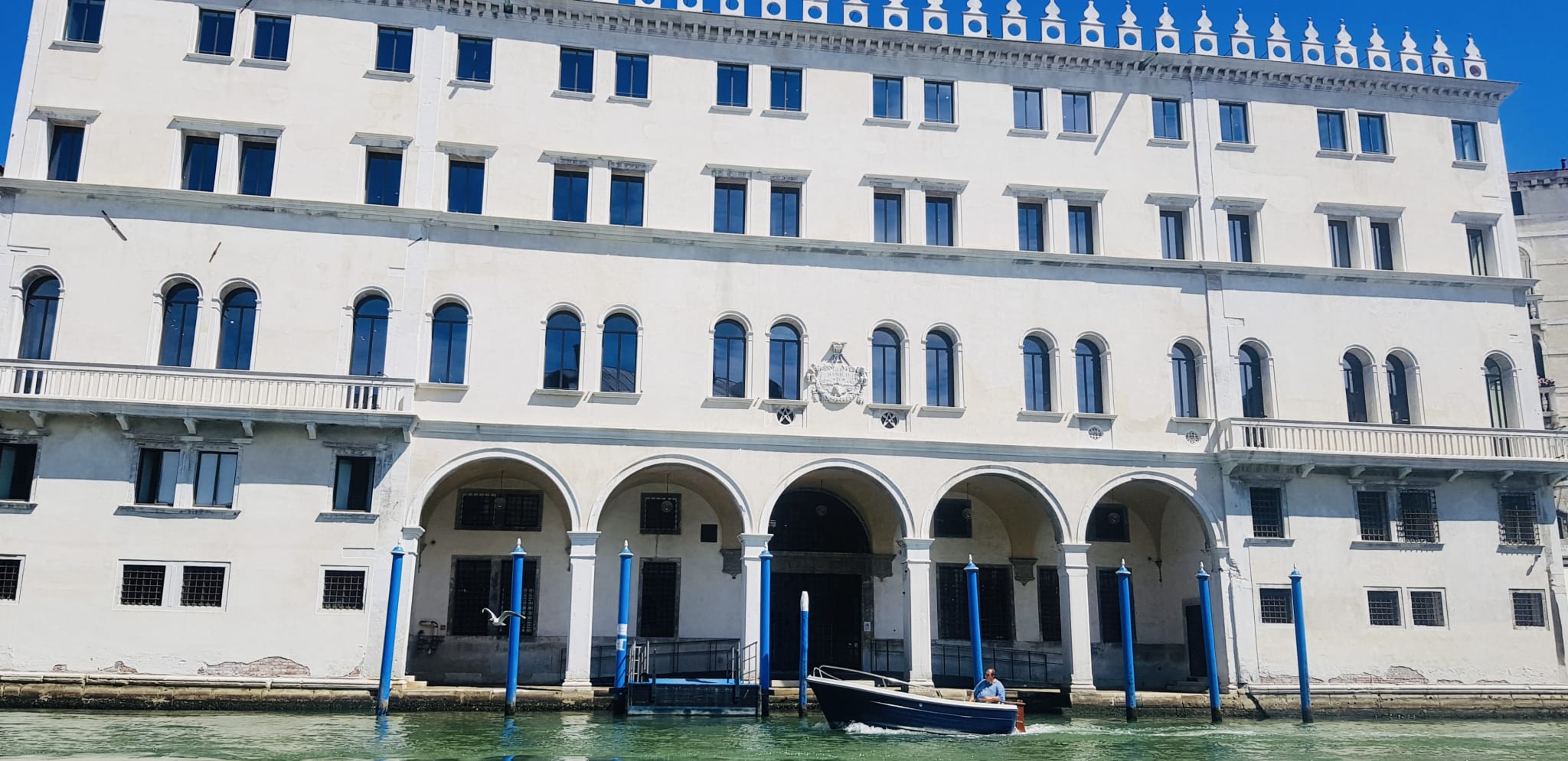 A small boat with a person inside navigates a canal in front of a large white building with arched windows and a decorative roofline, capturing the essence of Gay Venice. Known for its vibrant LGBTQ+ bars and welcoming atmosphere, this scene reflects the unique blend of culture and inclusivity that defines gay Venezia.