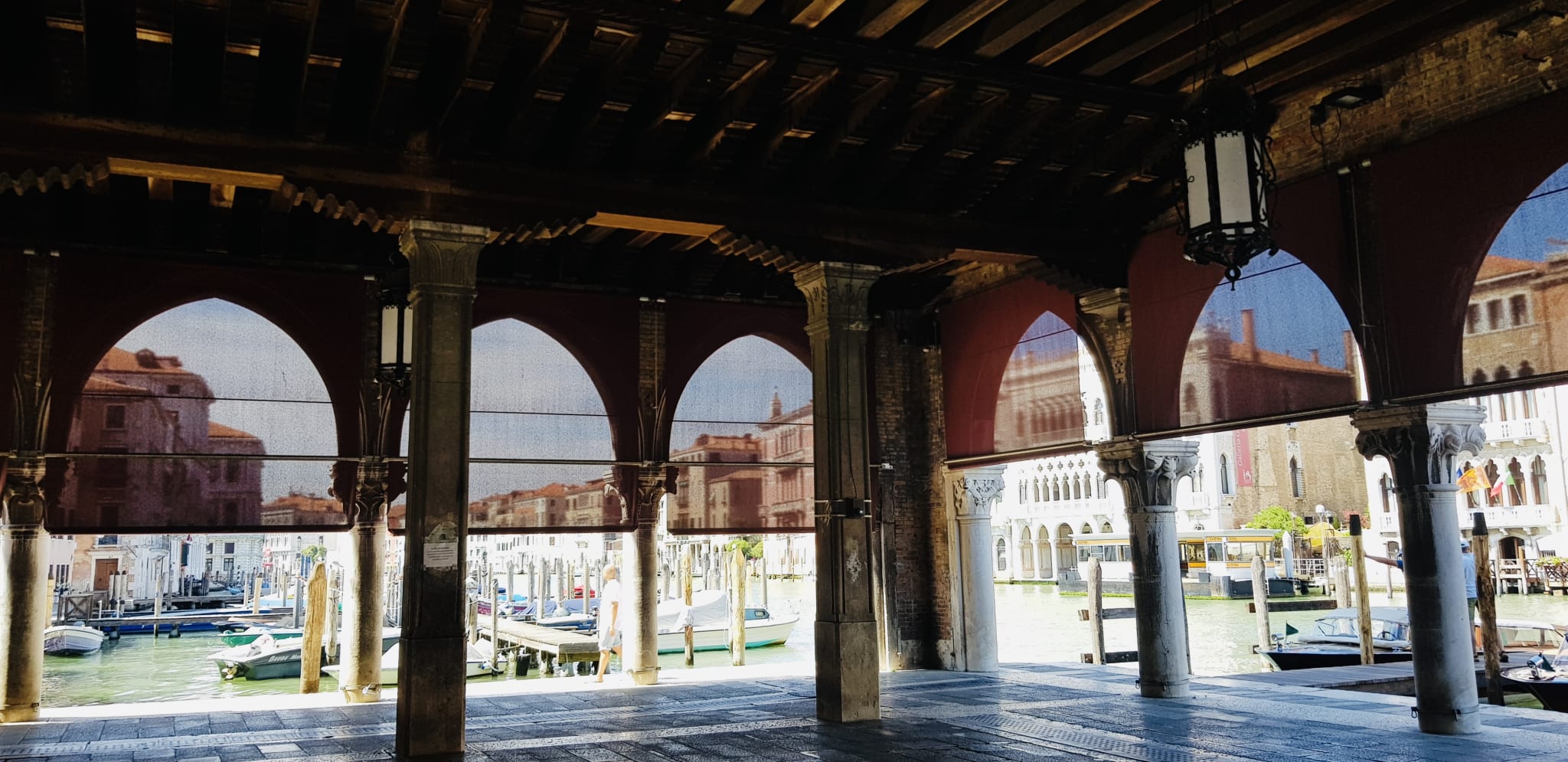 A covered walkway with arched openings overlooks a canal, where boats glide past historic buildings, encapsulating the vibrant spirit of Gay Venice. This enchanting city embraces diversity, with LGBTQ+ bars that thrive and gay-friendly hotels that offer a warm welcome to all travelers, making it an inviting destination for everyone.