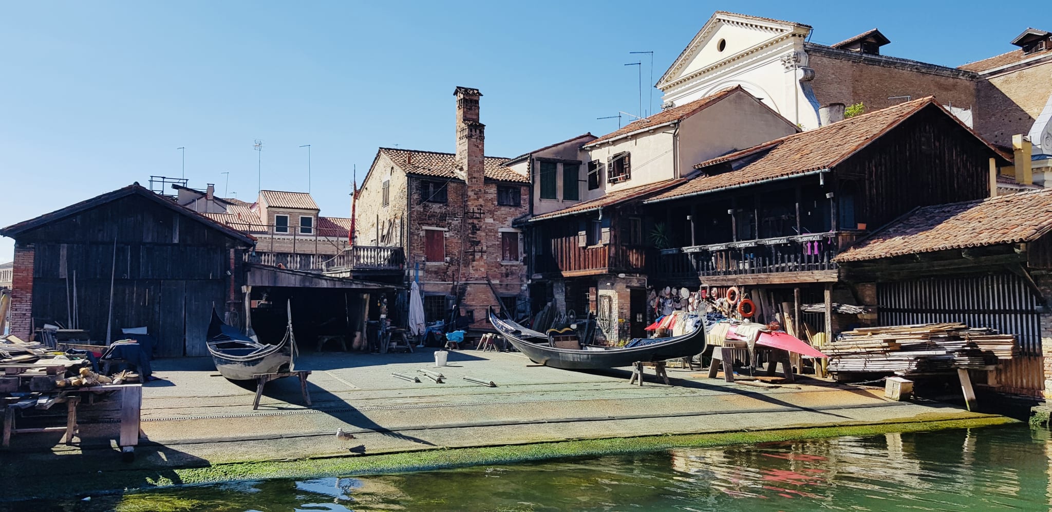 A charming boatyard with gondolas rests near the water, framed by wooden buildings under a clear sky—a picturesque setting for those exploring Venice.