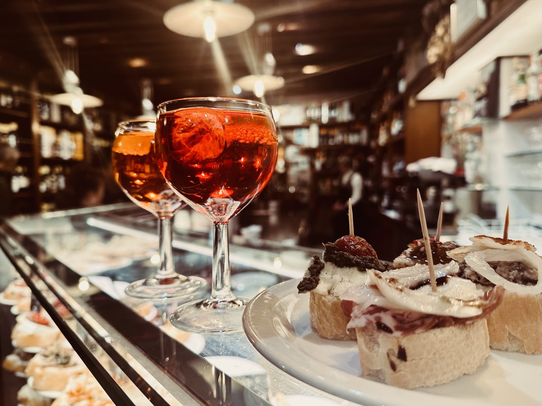 Two glasses of red aperitif rest gracefully on the bar, accompanied by a plate of authentic bites. The dimly lit restaurant captures the romantic charm and allure you'd expect on a 2-day Venice itinerary, offering an experience as rich and flavorful as Venetian culture itself. Perfect for unwinding after exploring canals or enjoying a gondola ride in this enchanting city.