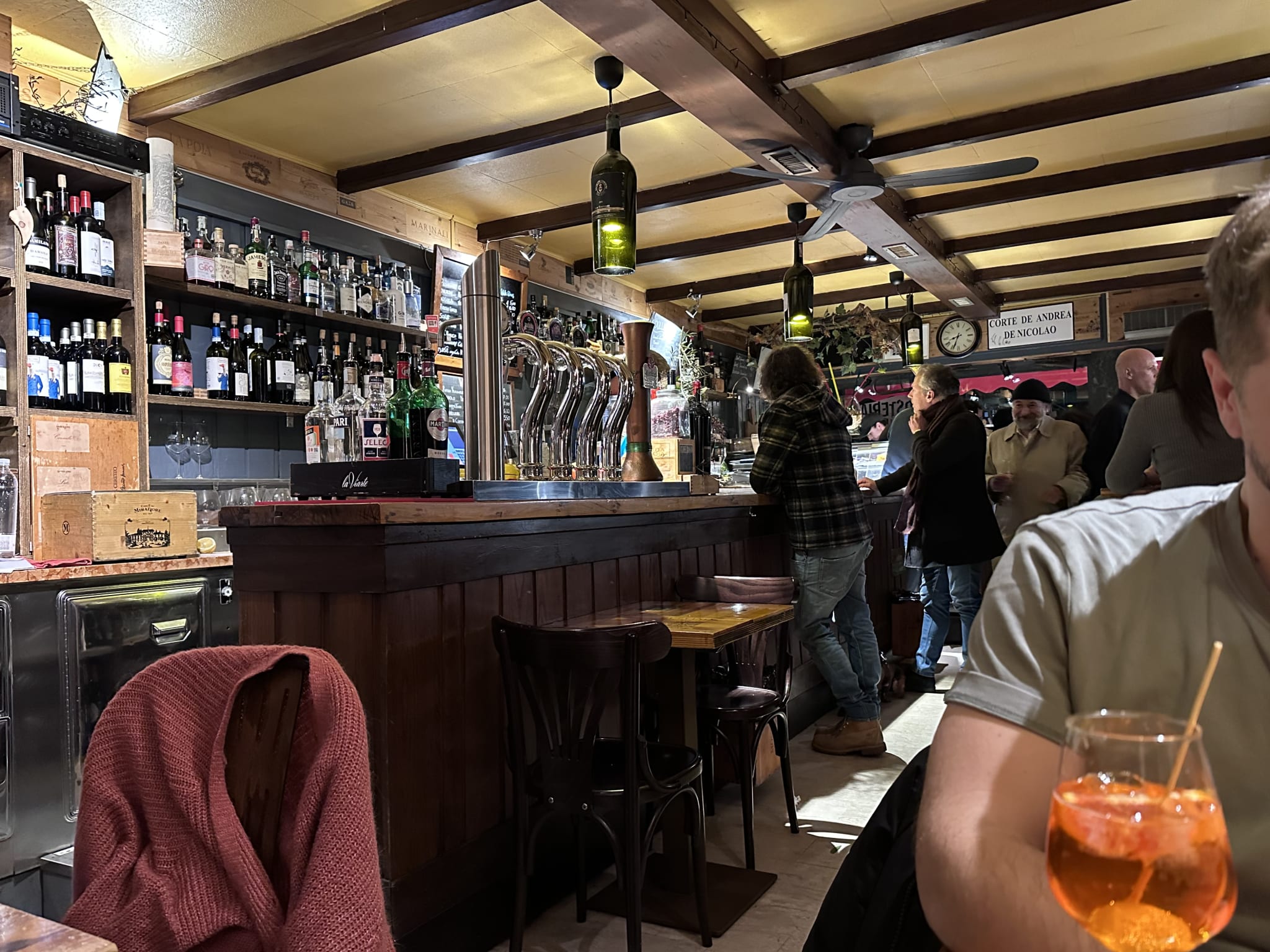 A cozy bar with wooden beams and shelves brimming with bottles captures Venetian history, perfect for a 2 day Venice itinerary. Patrons enjoy authentic bites, standing or seated at the counter. In the foreground, someone holds an orange drink reminiscent of an Aperol spritz—an iconic must-have as you explore cultural gems and culinary delights on your Venice adventure.
