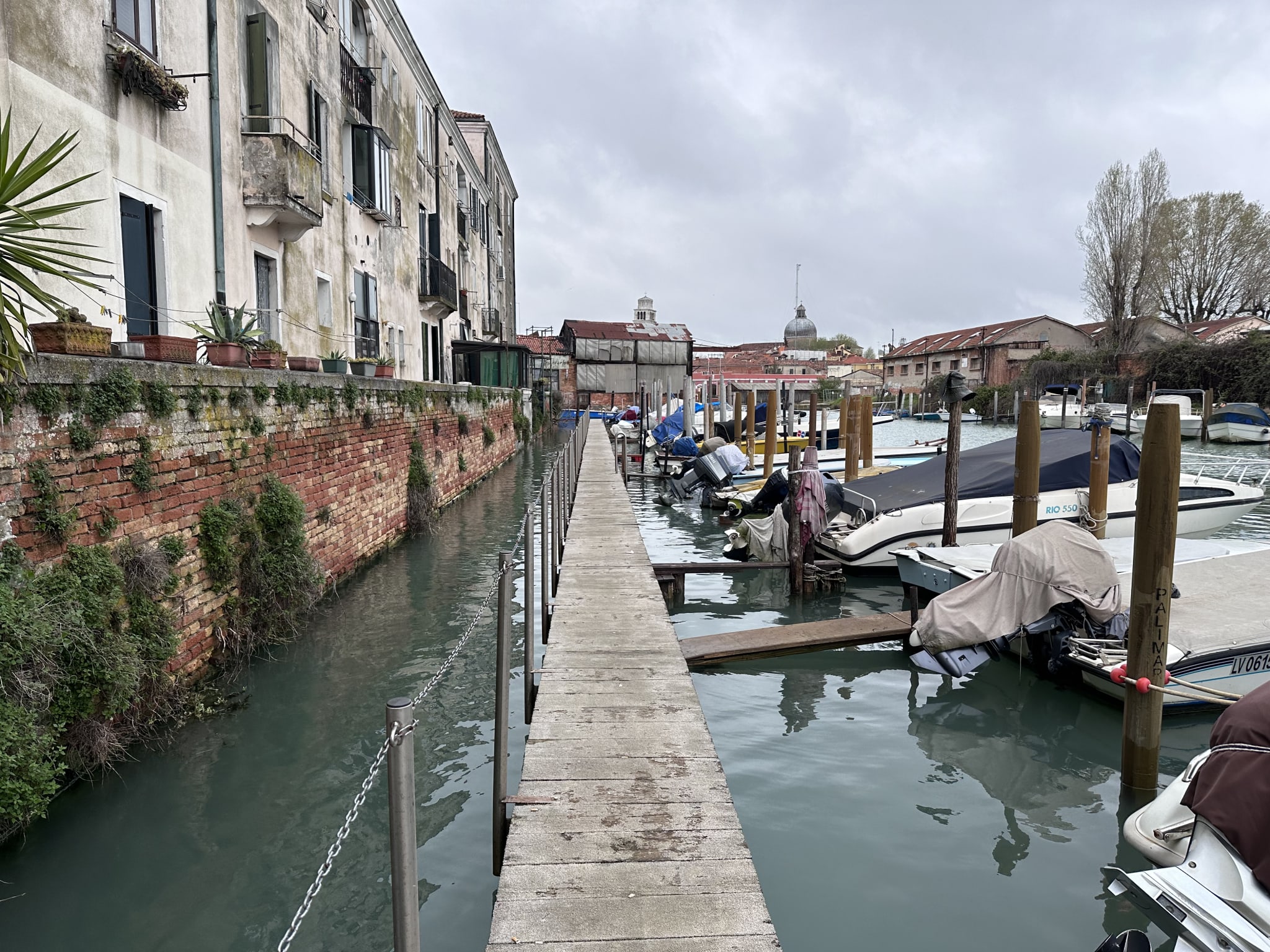 Narrow walkway hugging a serene canal where boats are docked on the right, with an ancient Venetian building heavy with history on the left, all under a moody cloudy sky. Perfect for a 2 day Venice itinerary, this scene captures the timeless charm and exploration potential of Venice's hidden paths and historical architecture.