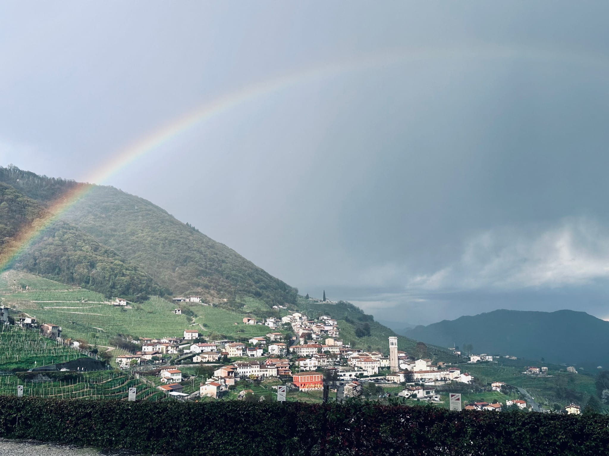 A rainbow elegantly curves over the quaint hillside village with lush green fields, charming houses, and a distant church tower under a cloudy sky. This serene scene echoes the enchanting canals and rich history found on a 2 day Venice itinerary—a perfect exploration of Venice's hidden gems and iconic landmarks for travelers seeking an unforgettable experience.