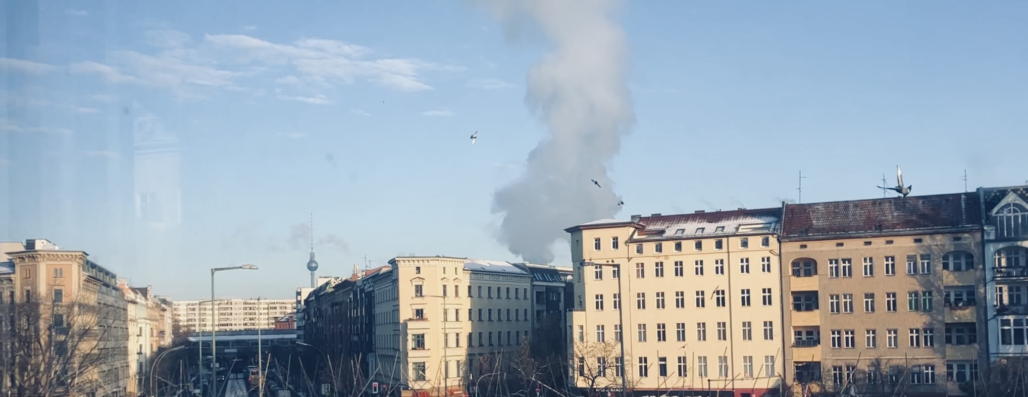 A view from Kreuzberg, Berlin, showing traditional buildings and a train bridge with a plume of smoke rising in the background, while the iconic TV Tower is visible in the distance, hinting at a vibrant yet calm cityscape.
