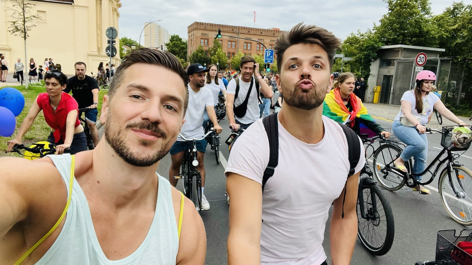 Two men in the foreground take a selfie at a fun cycling event, capturing memories of their adventures. One wears a white tank top, while the other sports a white t-shirt and makes a playful face. Behind them, cyclists ride on, including one with a pride flag and another in a pink helmet. Trees and buildings frame this lively scene.