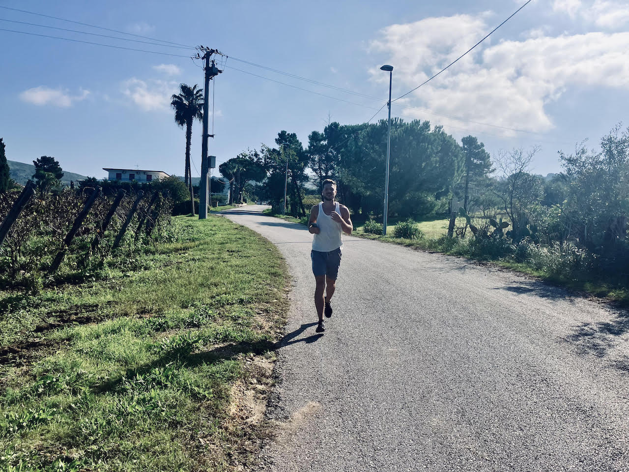 A person jogging on a sunny rural road lined with greenery and trees, symbolizing holiday adventures. The runner wears a white tank top and shorts, passing a lamp post and tall palm tree. Hills and a distant house under a partly cloudy sky complete this picturesque scene.