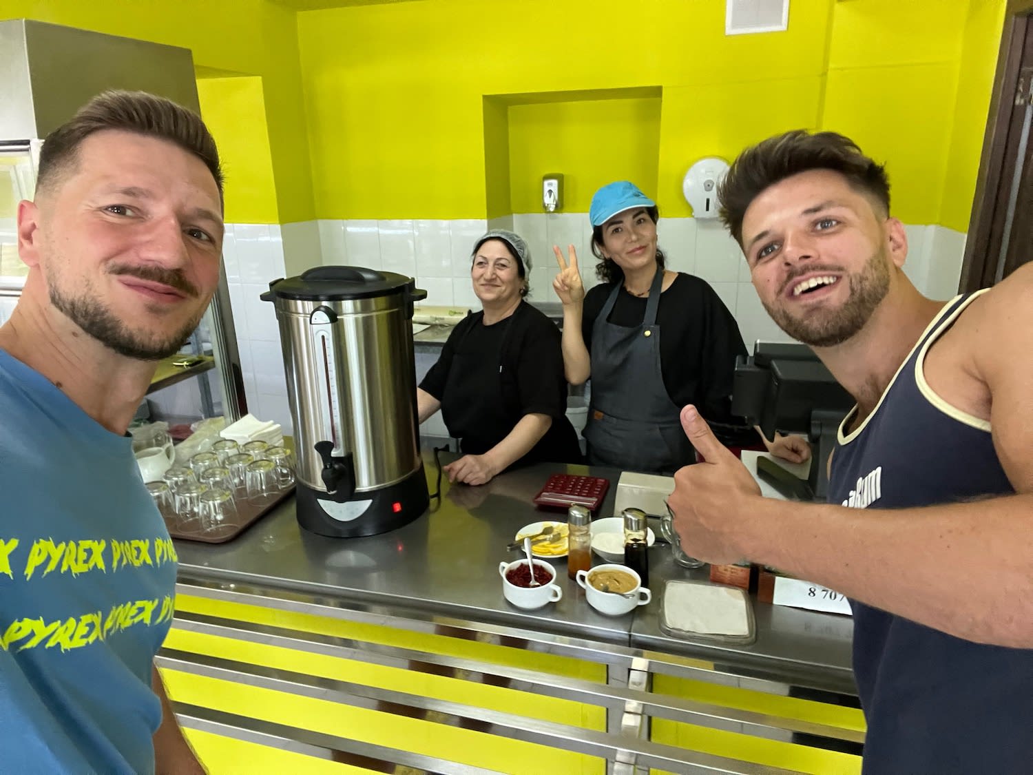 Two tourists giving thumbs up inside a traditional Kazakhstani canteen with friendly local staff in Almaty, capturing the warm hospitality.